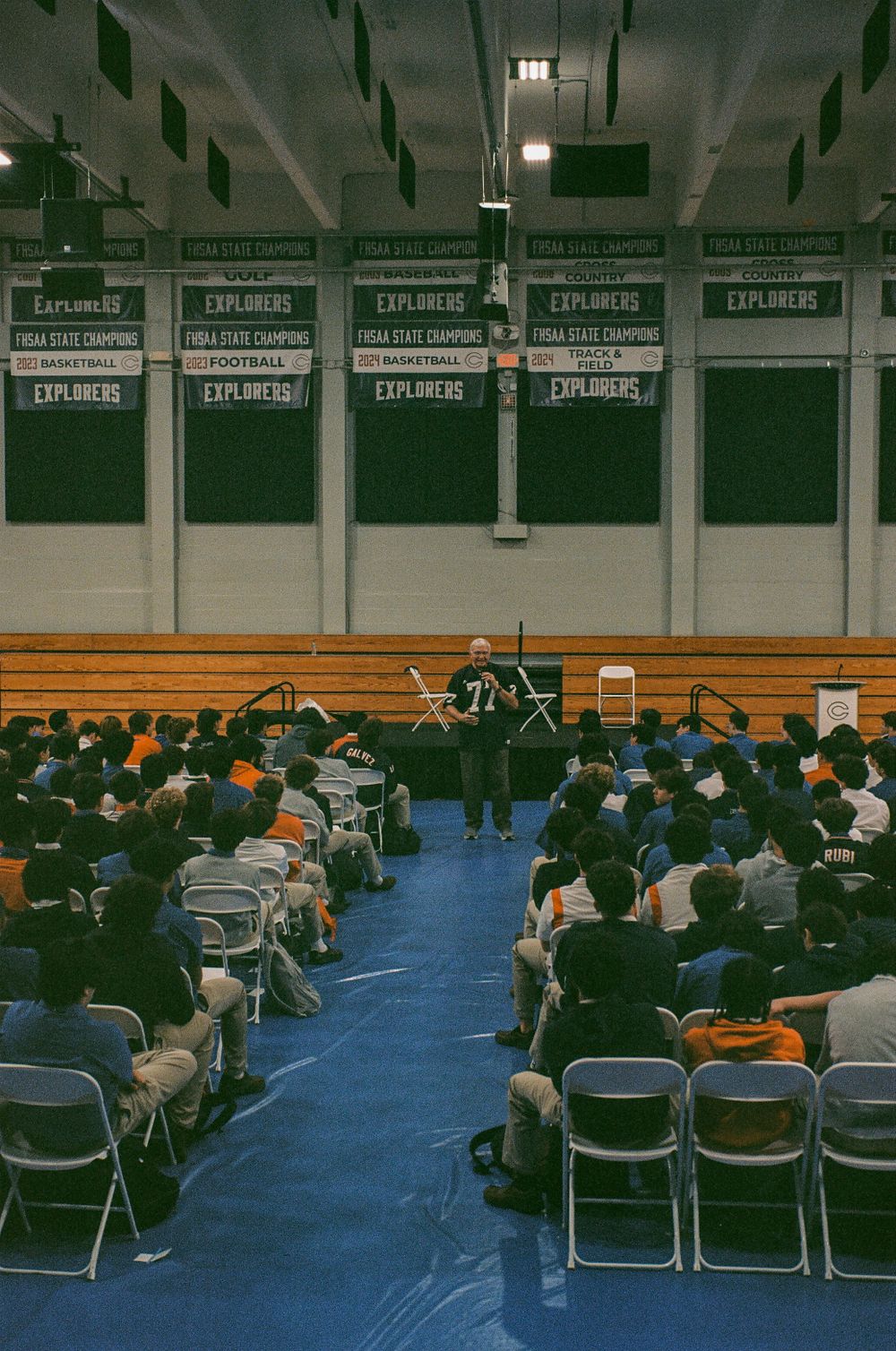 A large audience seated in folding chairs watches a presentation in a gymnasium with banners.
