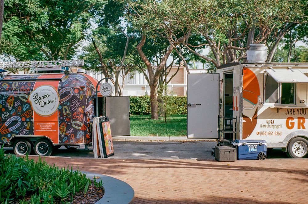 Two food trucks parked along a tree-lined path in an outdoor venue.