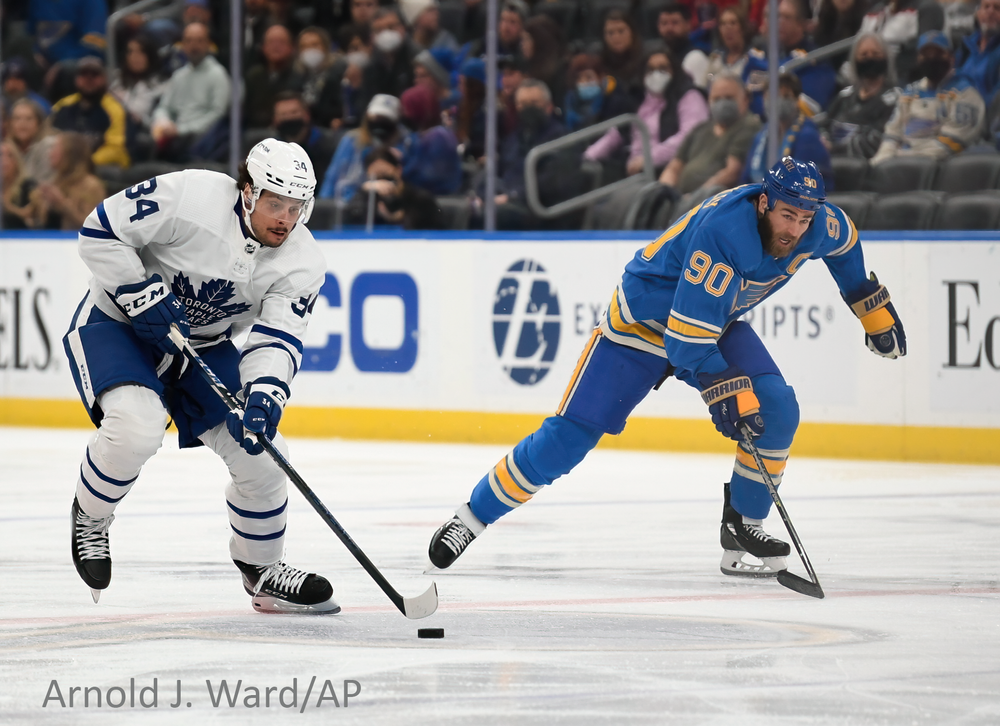 Two hockey players, one in white and one in blue uniforms, battle for control of the puck during an NHL game.