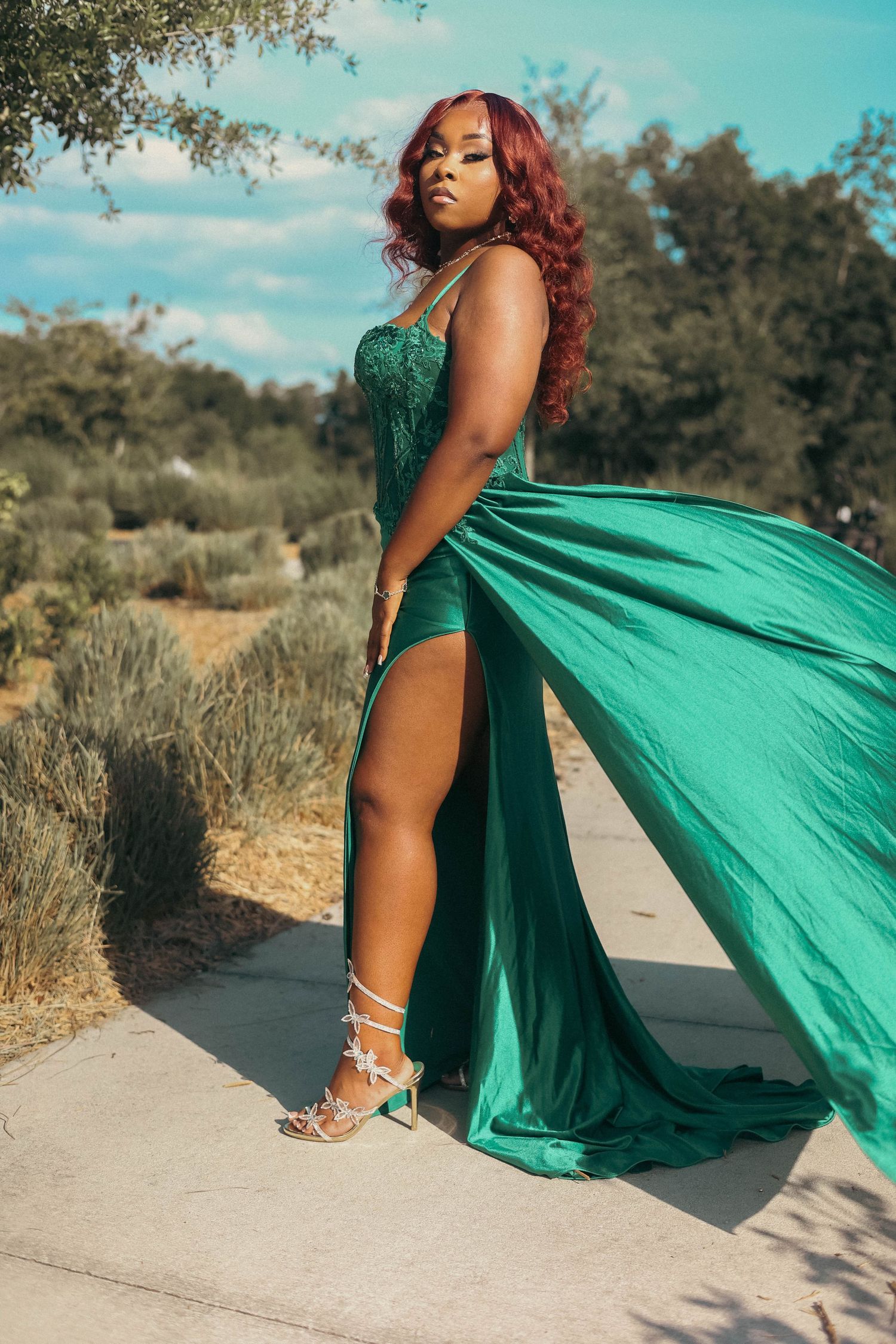 Woman in elegant green satin gown with high slit posing outdoors on pathway with flowing fabric in breeze.