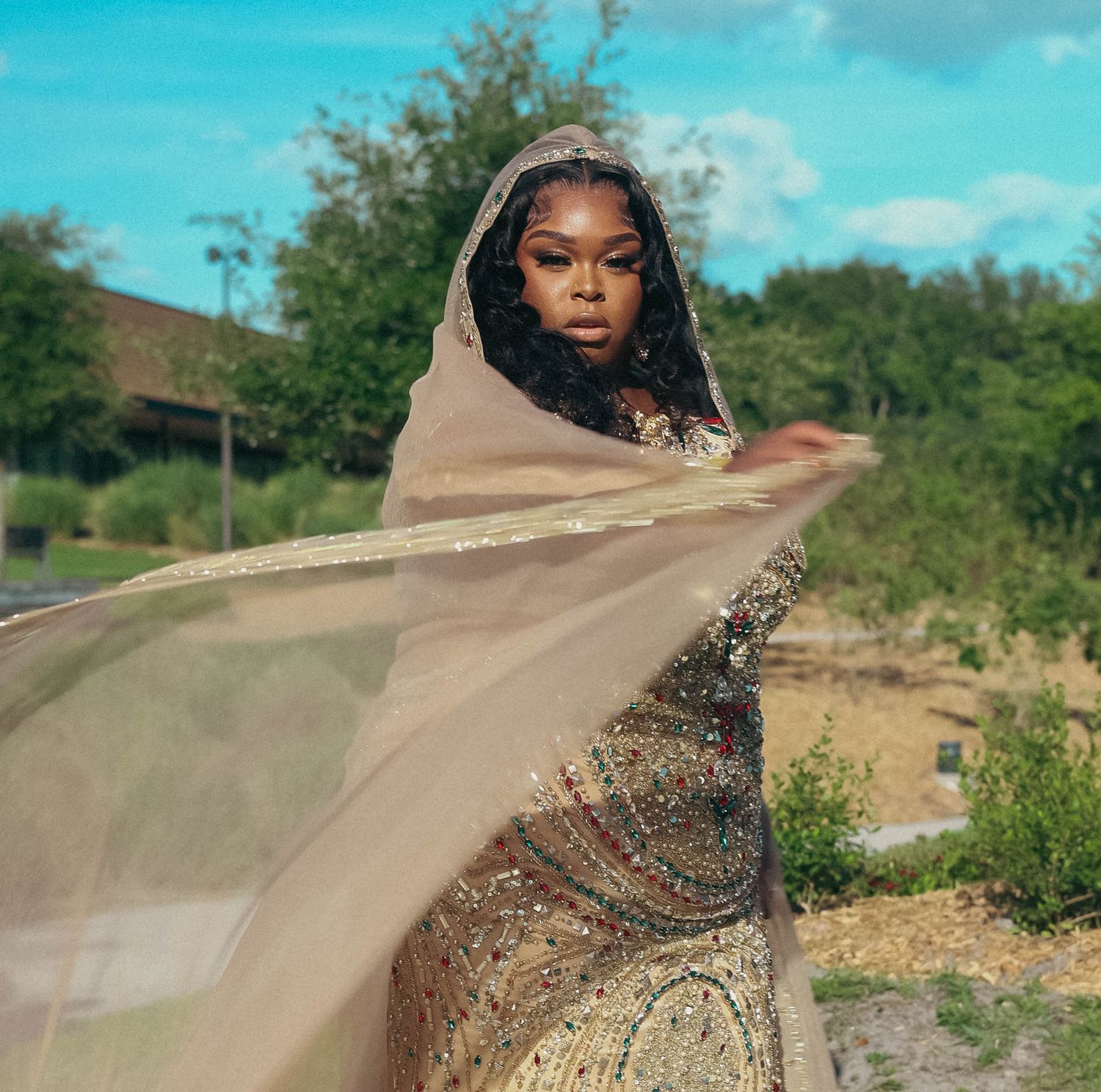 Woman dramatically spreading a sheer gold cape while wearing a beaded gown outdoors.