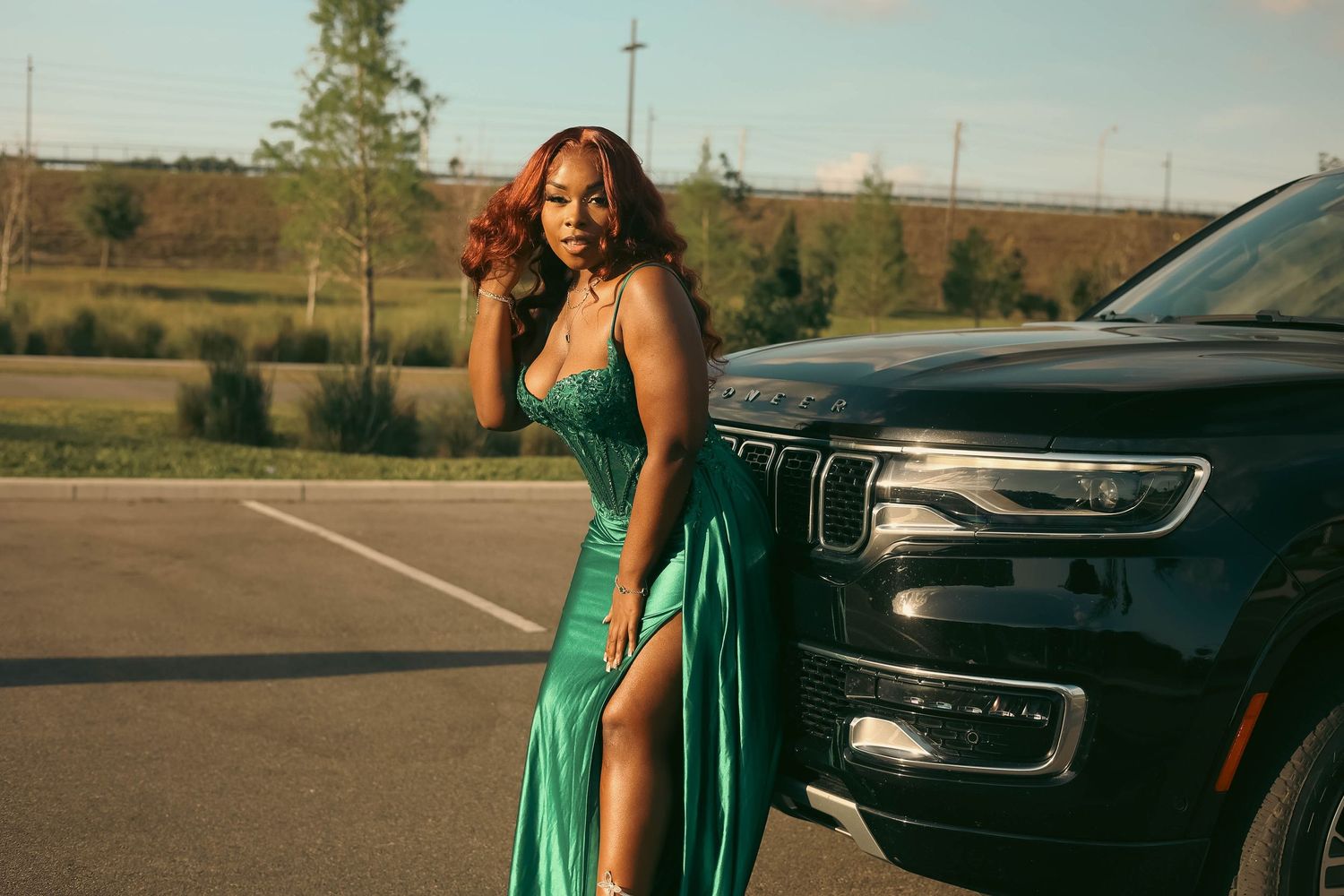 Woman in green satin formal dress with high slit posing beside black SUV in parking lot at sunset.
