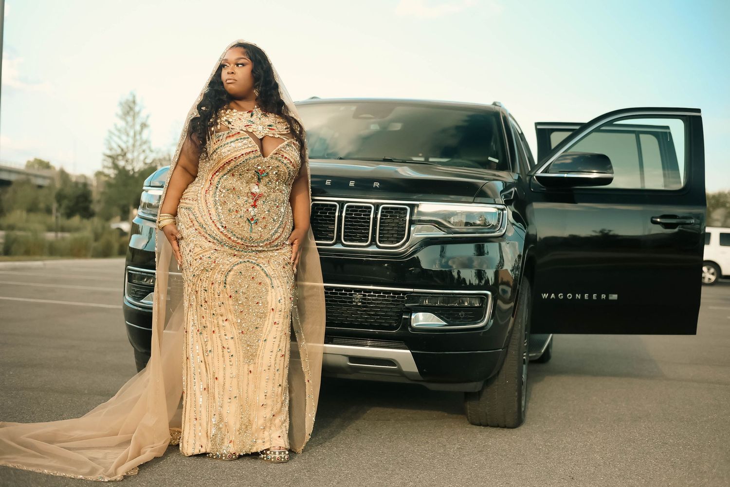 Woman in gold beaded gown with cape posing beside a black Jeep SUV.