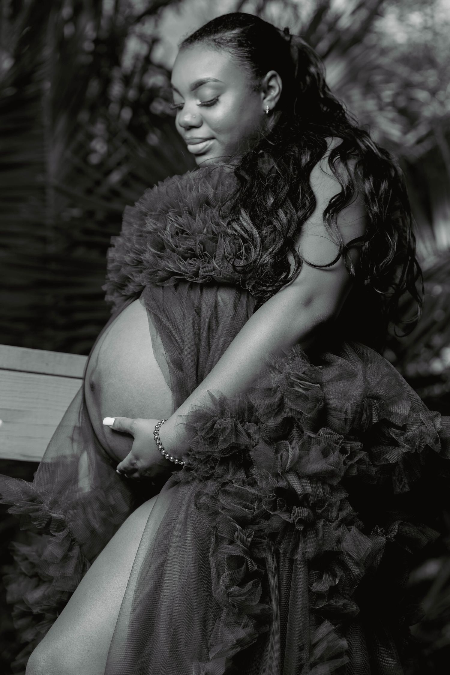 Black and white maternity portrait of a pregnant woman in a ruffled tulle gown seated outdoors.