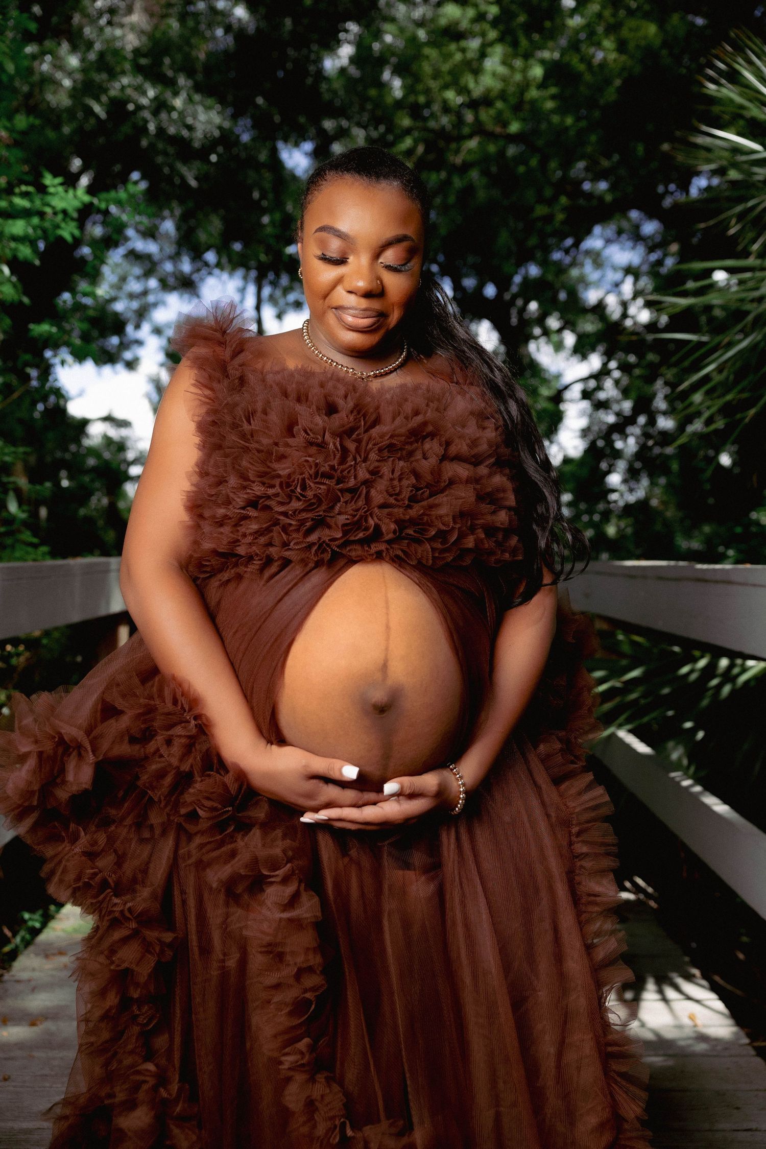 Elegant maternity portrait of a pregnant woman in a brown ruffled tulle gown cradling her baby bump.