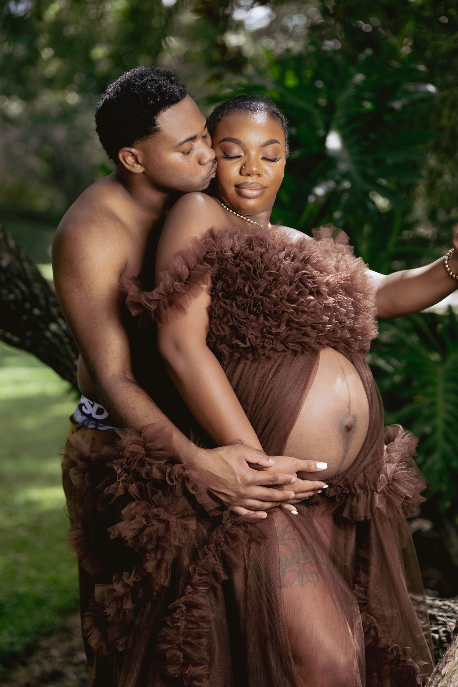 A couple sharing a tender maternity photo shoot outdoors, with man kissing woman in brown tulle gown.