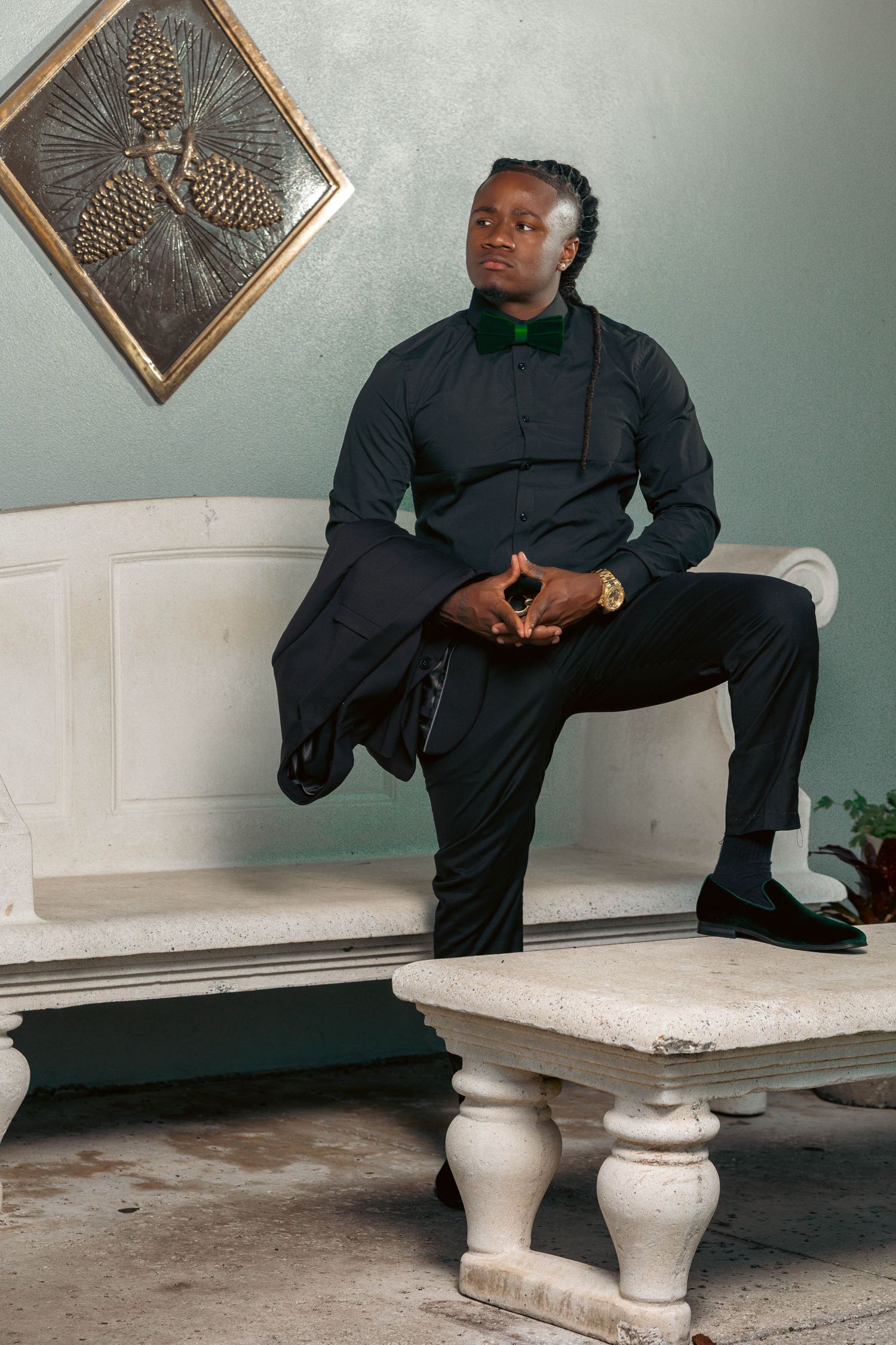 Well-dressed Black man in dark suit with green bow tie poses on white bench in elegant interior setting.