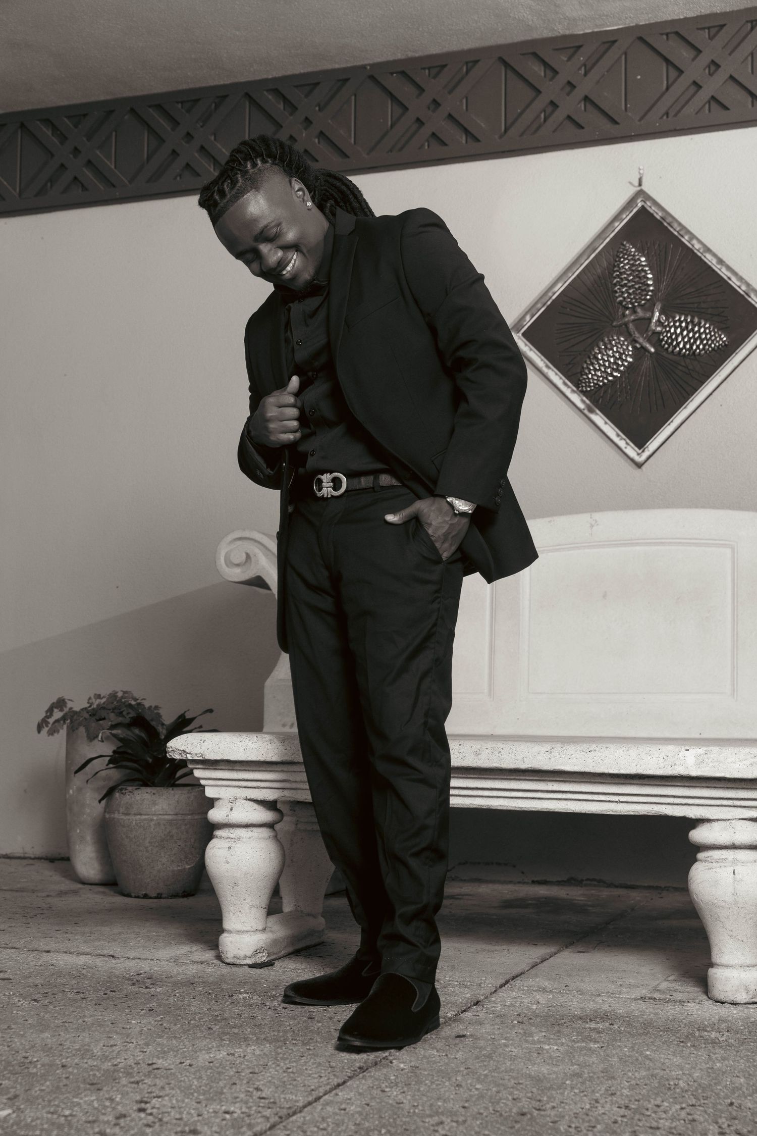 Stylish Black man in black suit laughs confidently beside white bench in black and white portrait photo.