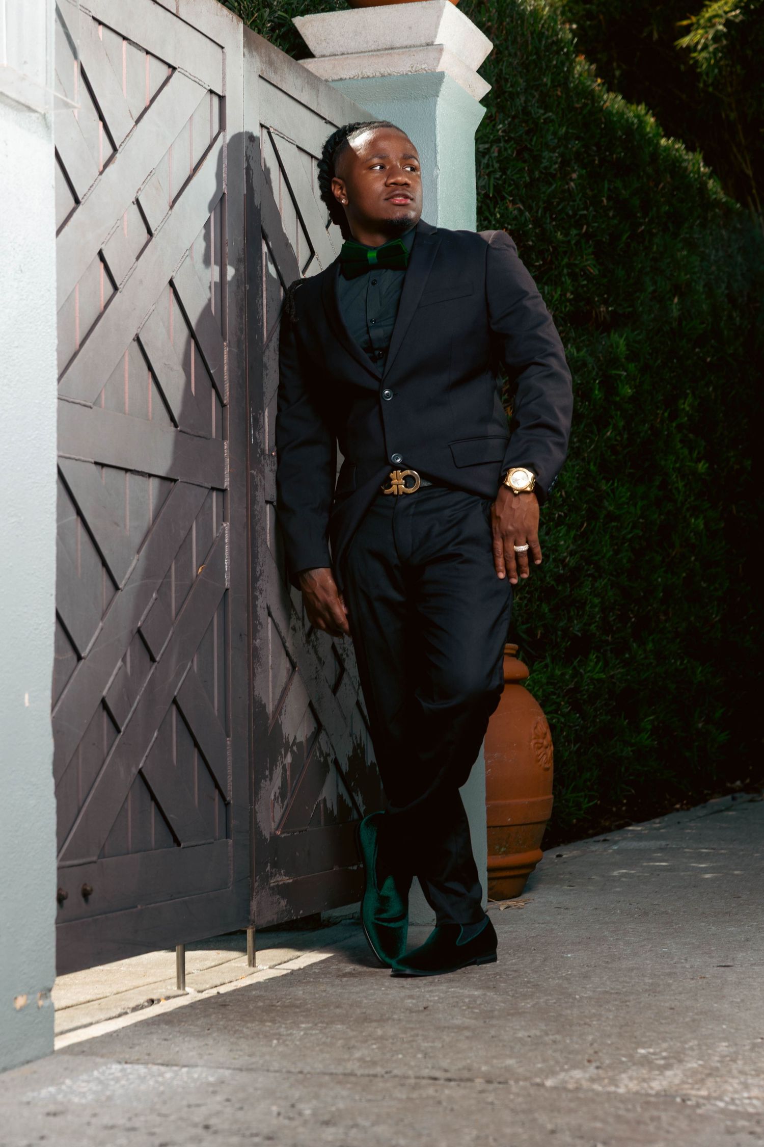 Dapper Black man in navy suit leans against decorative gray gate surrounded by lush green hedges outdoors.