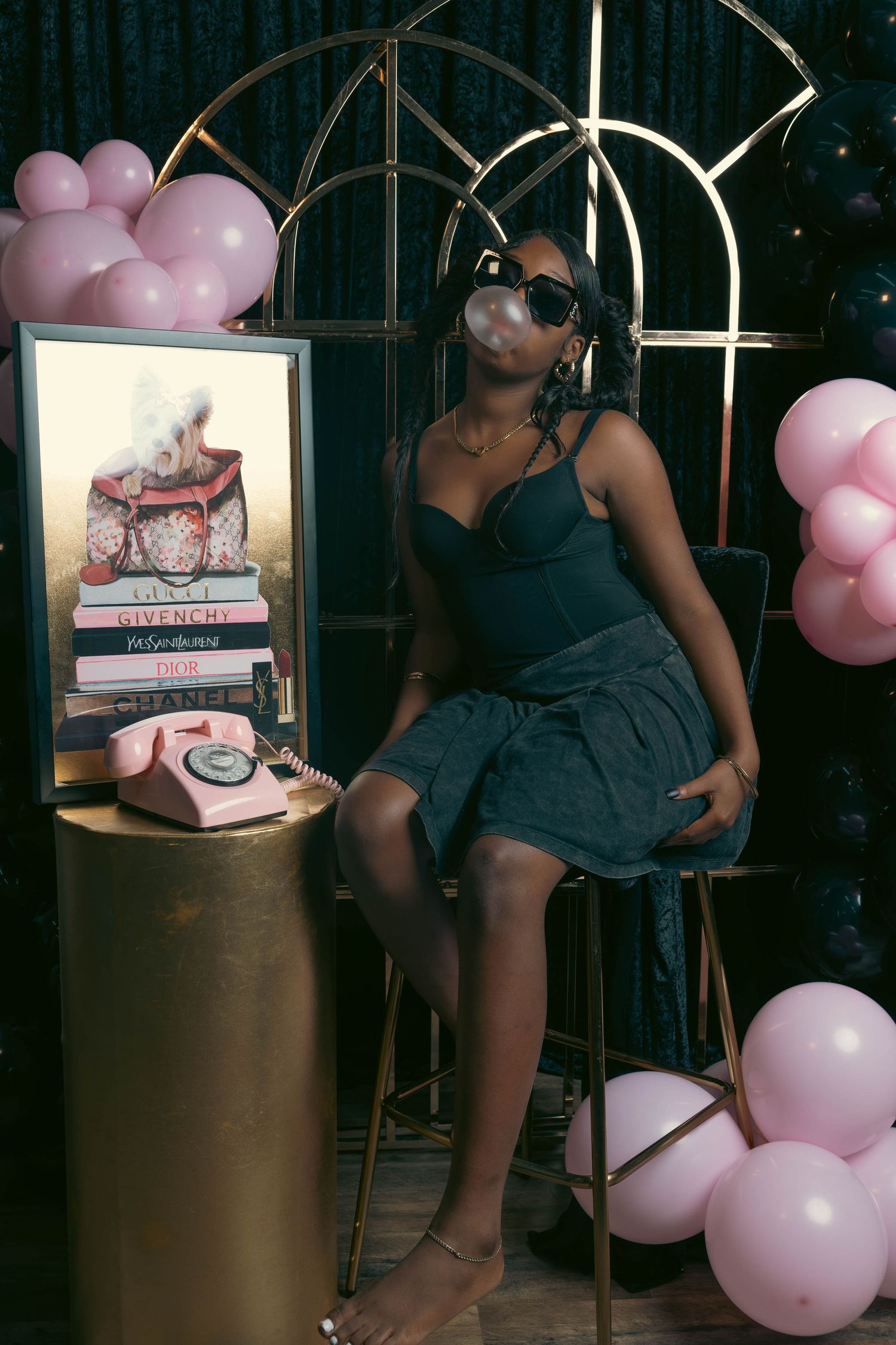Woman in black corset top blowing bubble gum surrounded by pink balloons in decorated studio.