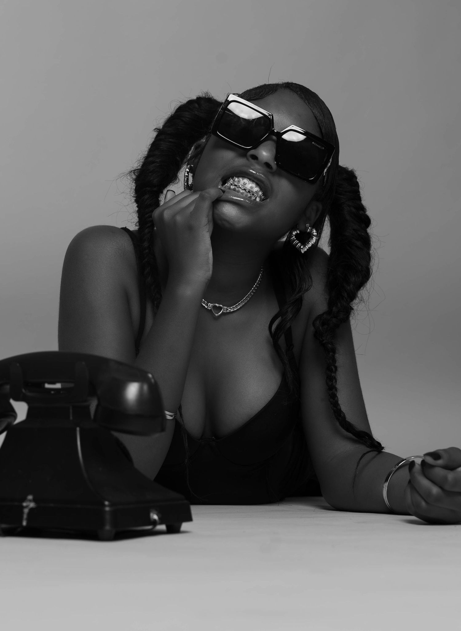 Black and white portrait of woman with braids and sunglasses posing beside vintage rotary phone.