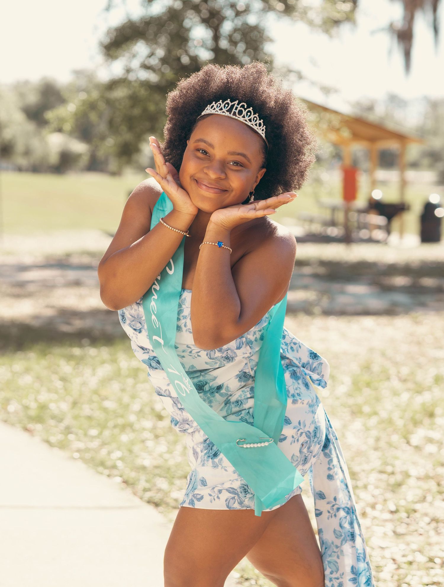 Young girl in floral dress and tiara with teal sash posing outdoors.