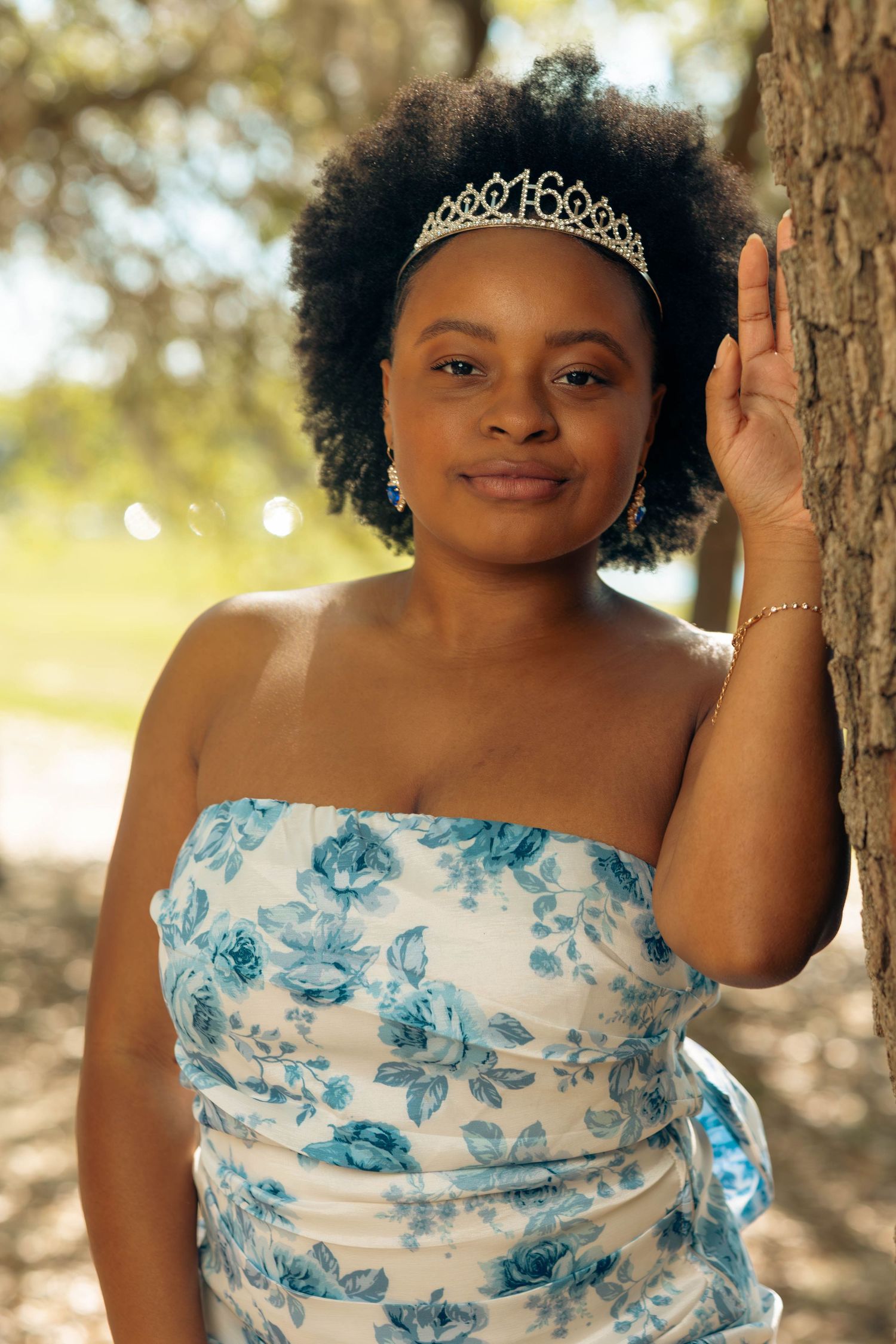 Teen girl wearing a sweet 16 tiara and floral strapless dress leaning against a tree.
