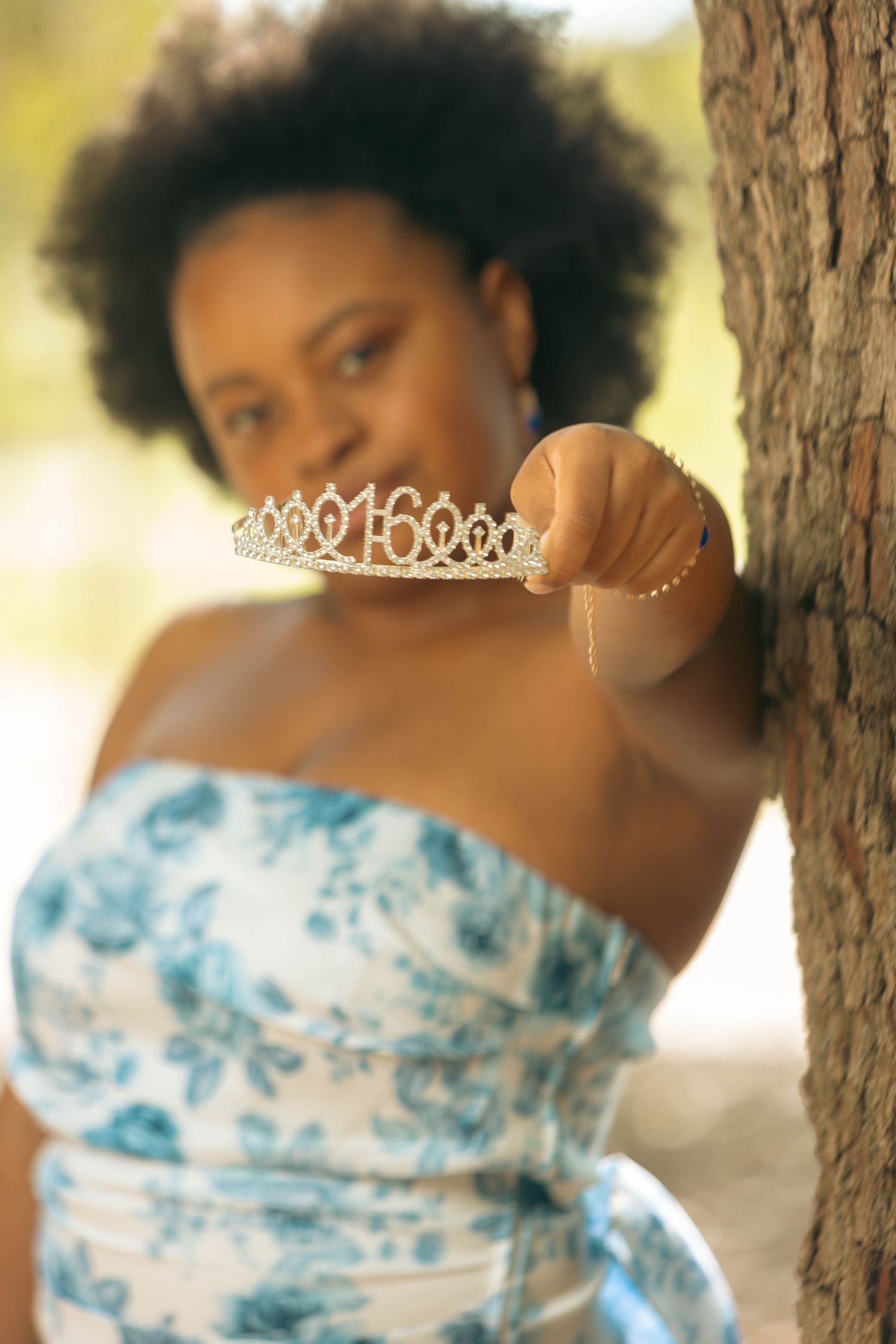 Teen girl holding a sweet 16 tiara toward camera while leaning against a tree.