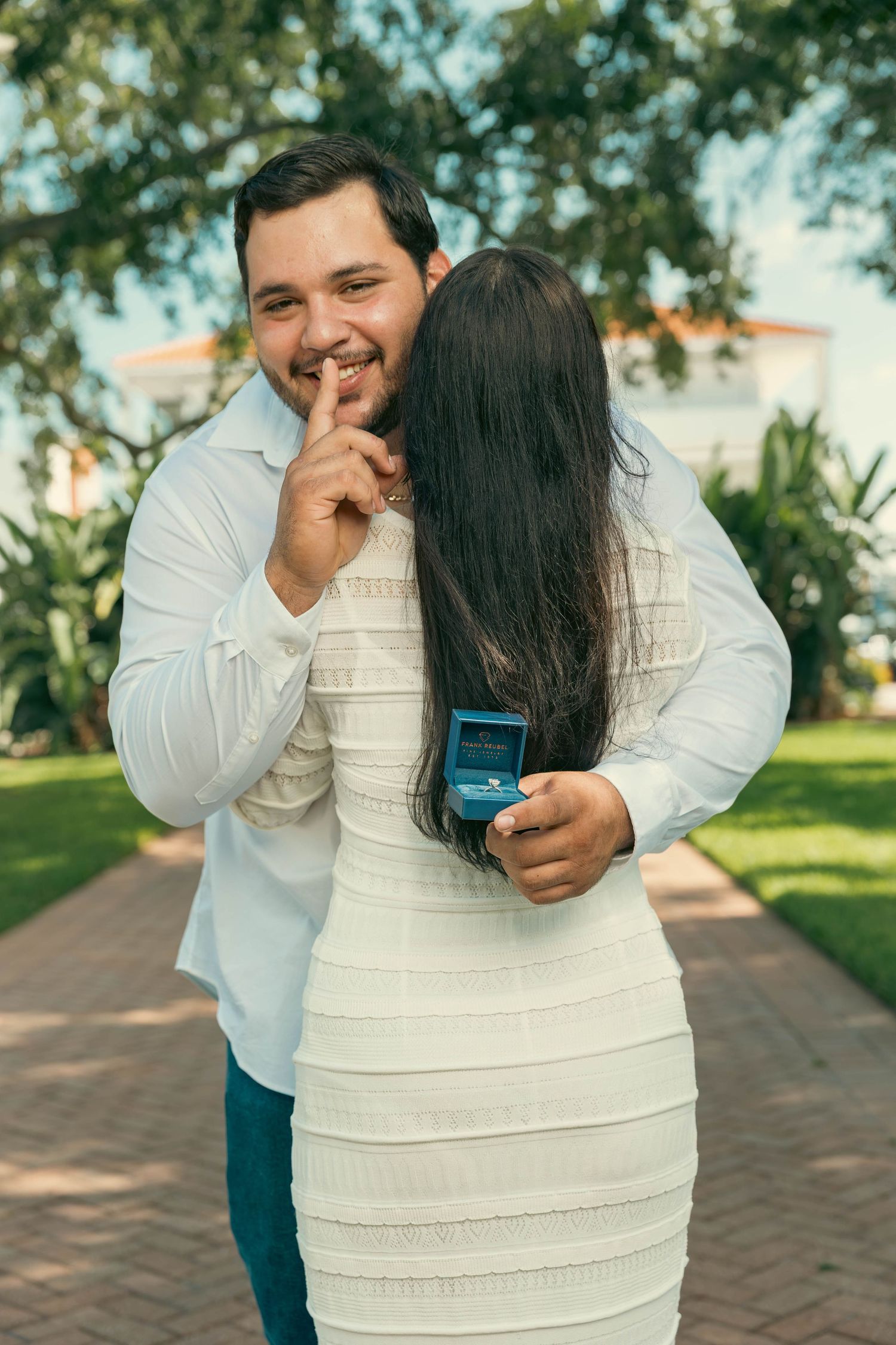 Smiling man holding engagement ring box behind woman in white dress in outdoor garden setting.