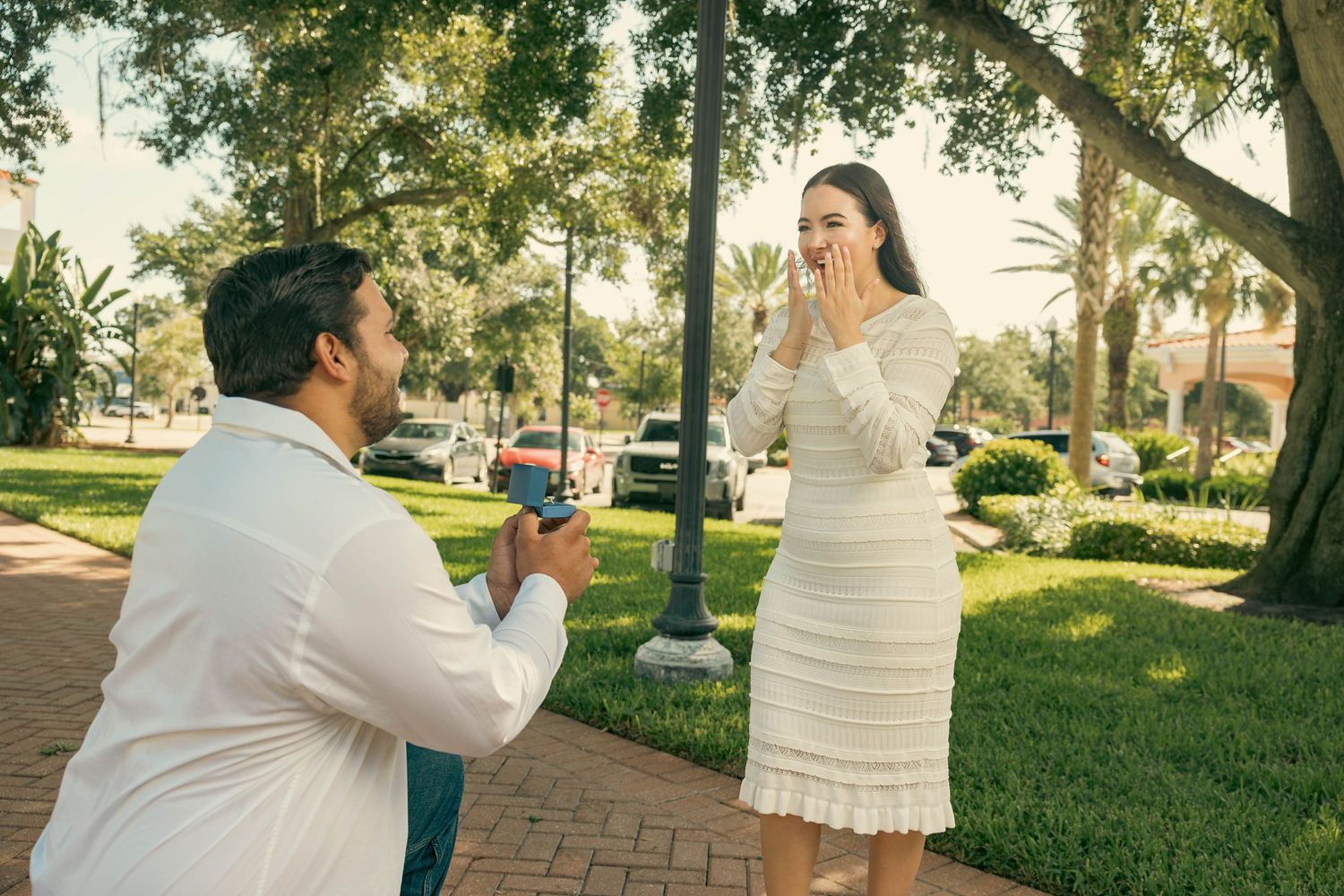 Man on one knee proposing to surprised woman in white dress outdoors in a park.