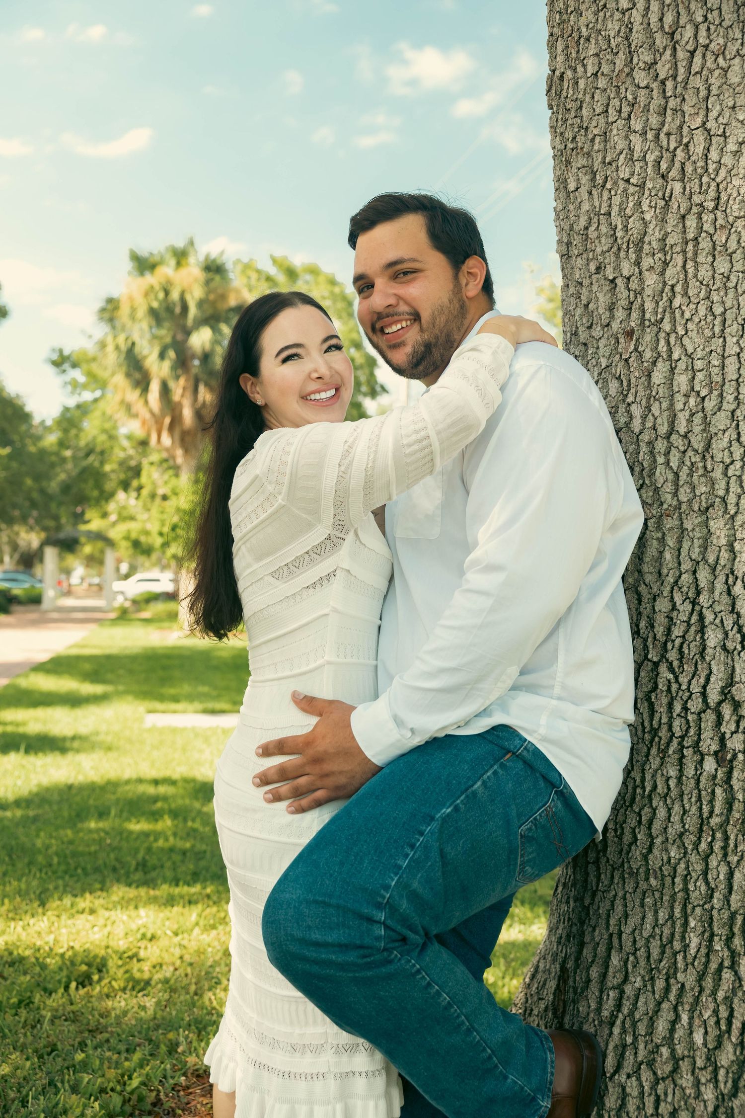 Happy couple in white outfits embracing and smiling by a tree in a sunny park.