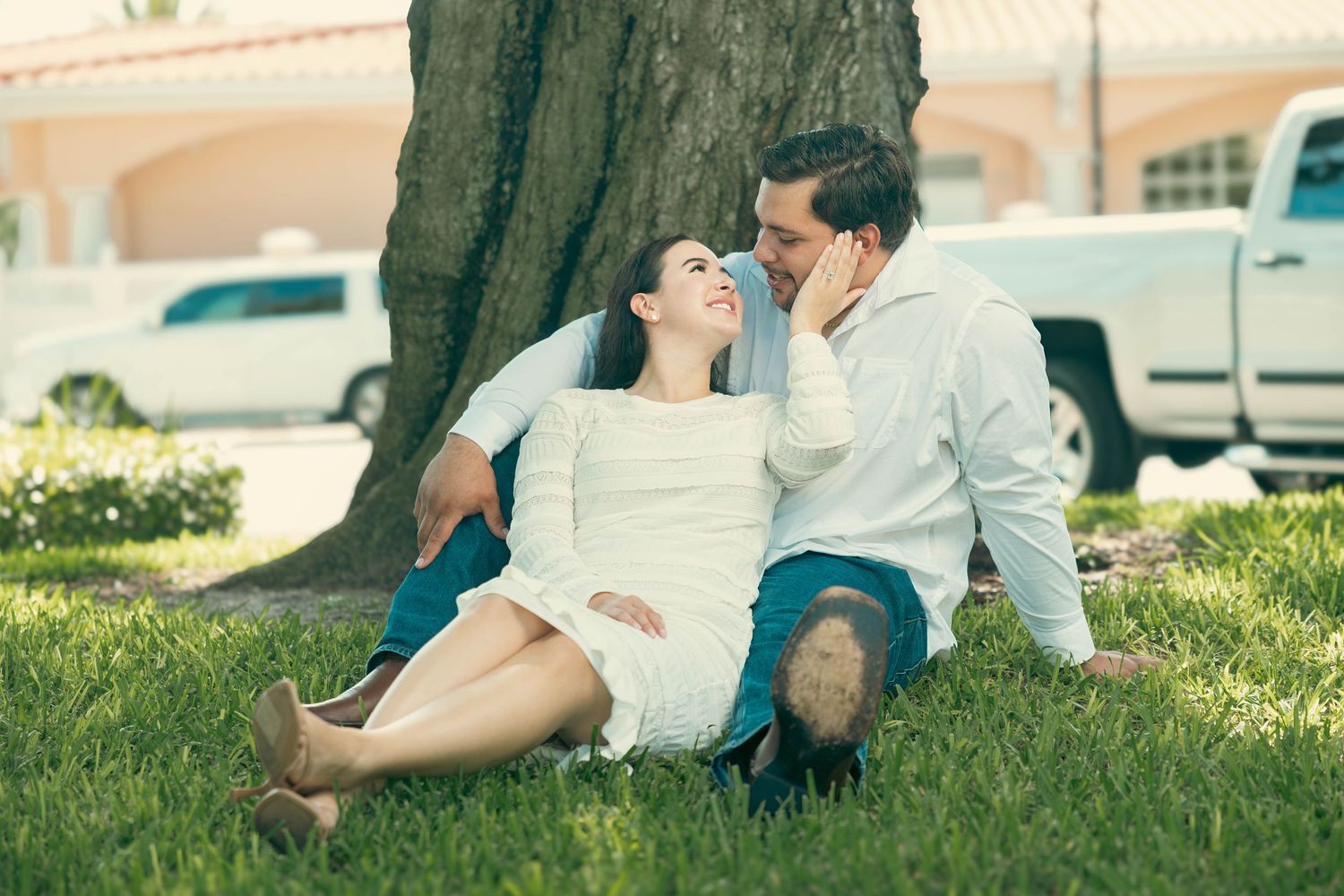 Romantic couple sitting together against a tree trunk on green grass outdoors.