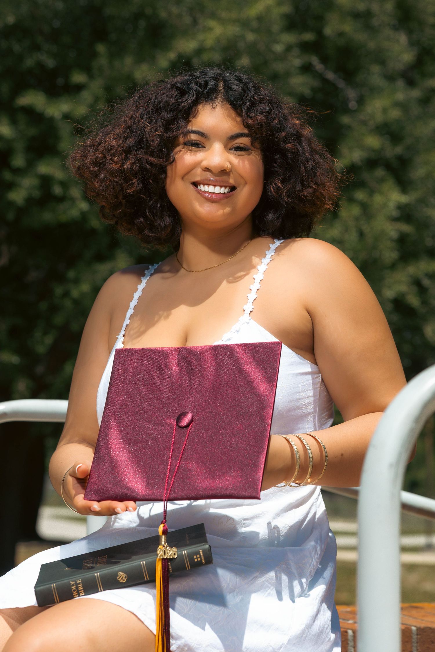 Smiling graduate in white dress holds a maroon cap and Holy Bible outdoors in sunlight.