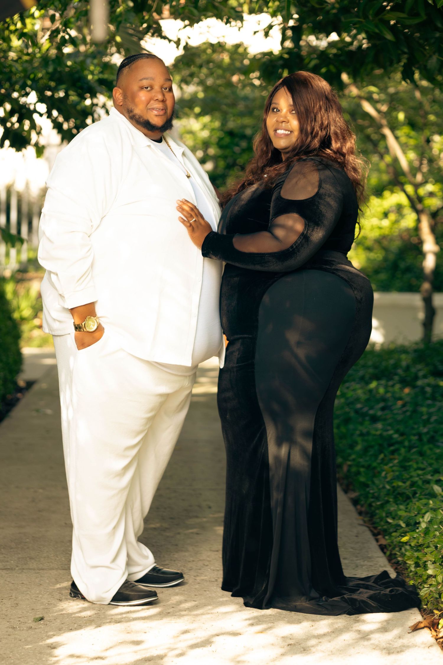 Couple poses outdoors man in white suit woman in elegant black mesh gown among green trees.