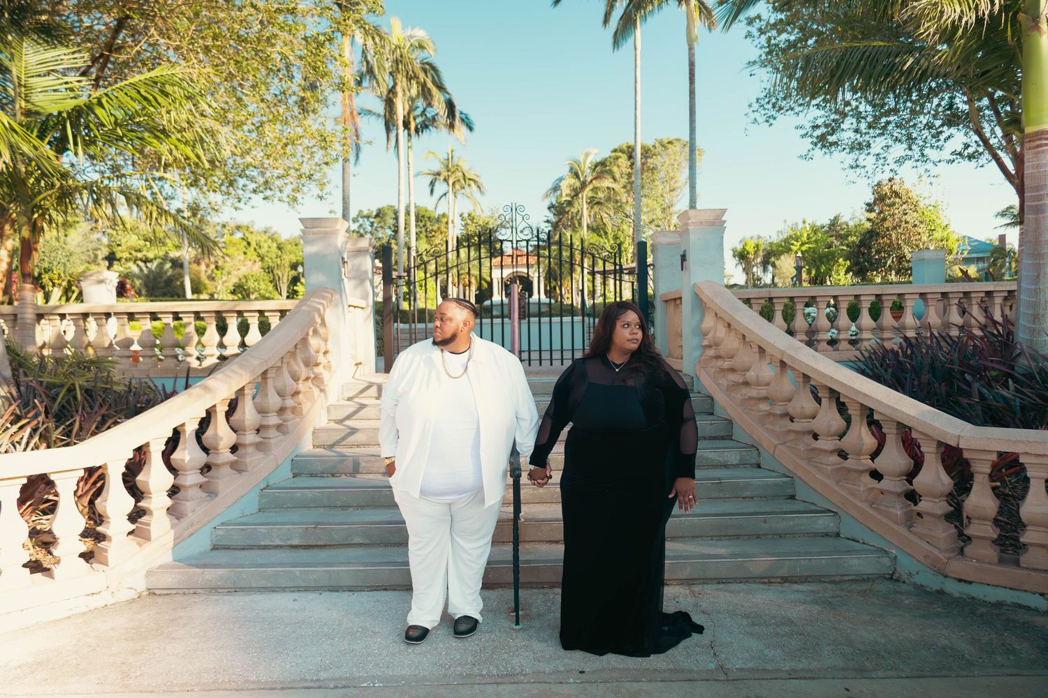 Couple walking down elegant outdoor staircase lined with white balusters surrounded by palm trees.