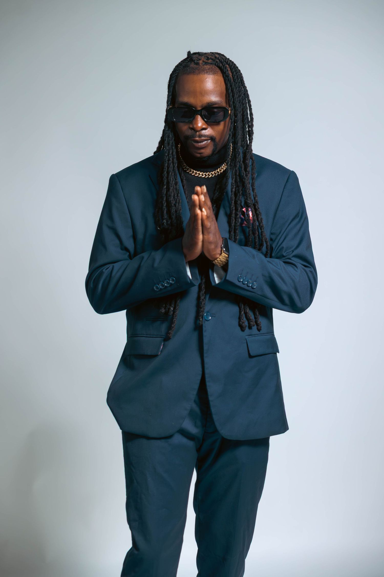 Stylish man in navy blue suit with long dreadlocks and sunglasses pressing palms together.