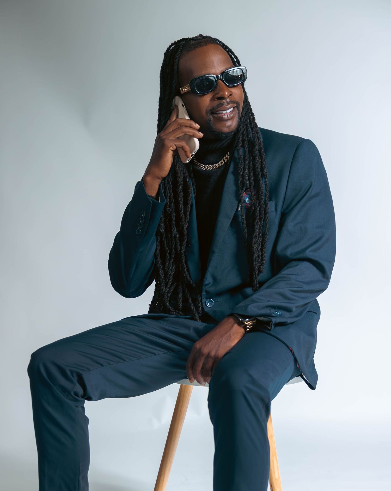 Man with long dreadlocks in teal suit sitting on stool talking on phone wearing sunglasses.