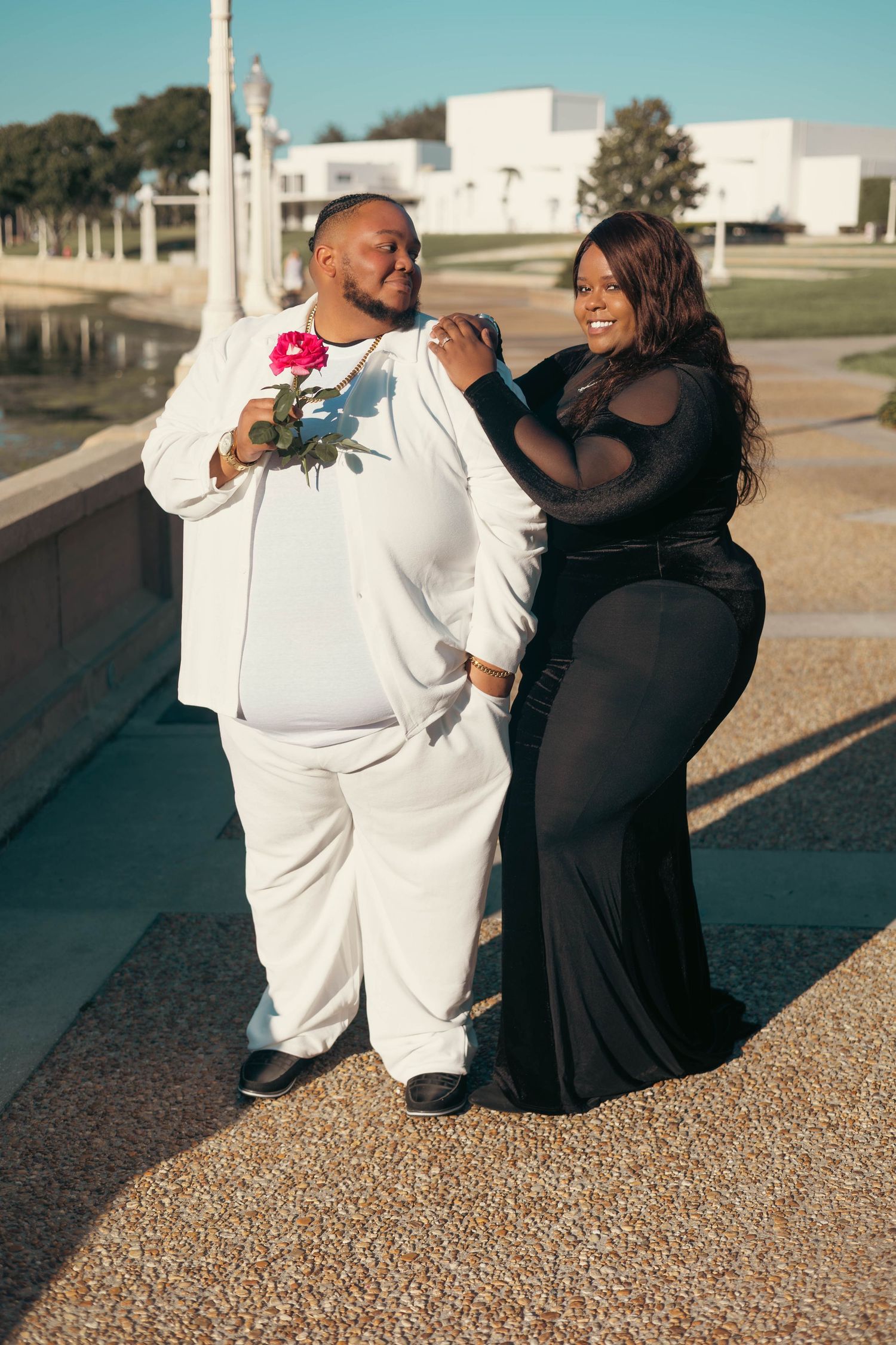 Couple poses outdoors man in white suit holding rose woman in black gown leaning on his shoulder.