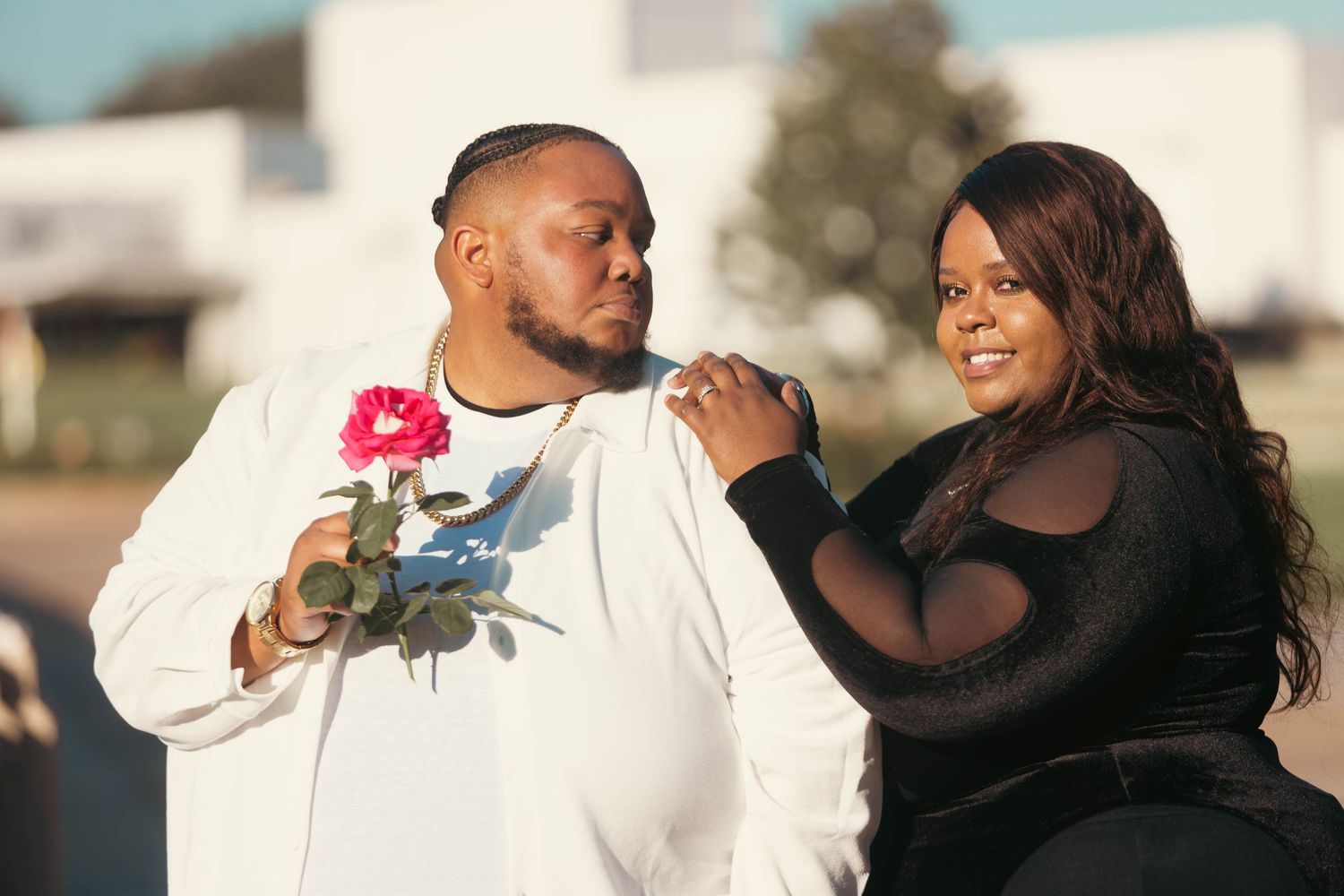 Man in white outfit holding pink rose stands beside woman in black outfit in an outdoor urban setting.
