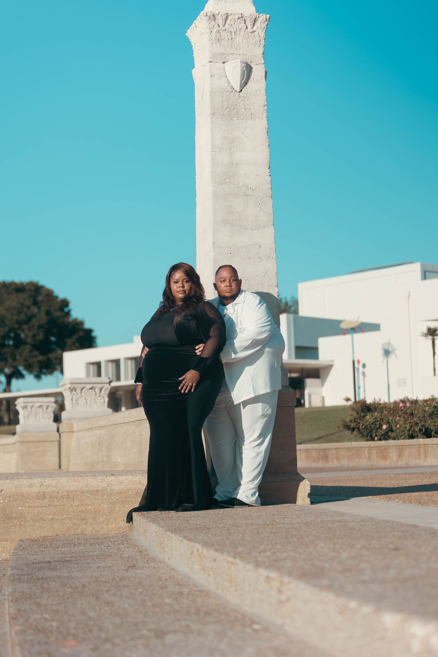 Elegant couple posing by a monument, woman in black gown and man in white suit.