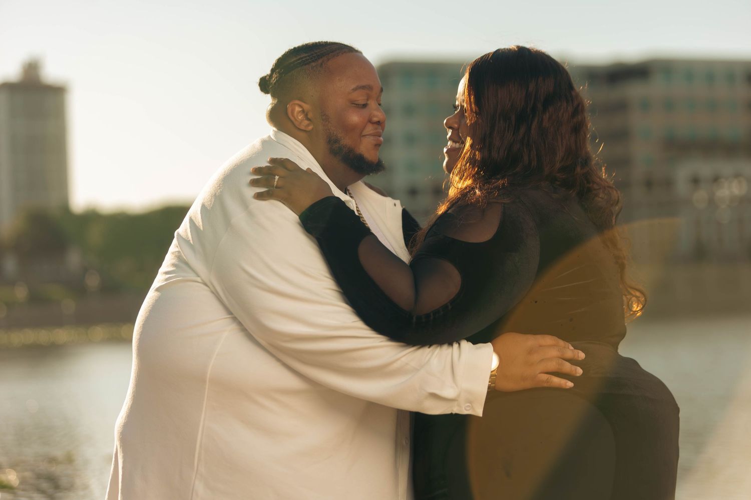 Couple sharing a warm embrace and smiling at each other with a sunlit waterfront in the background.