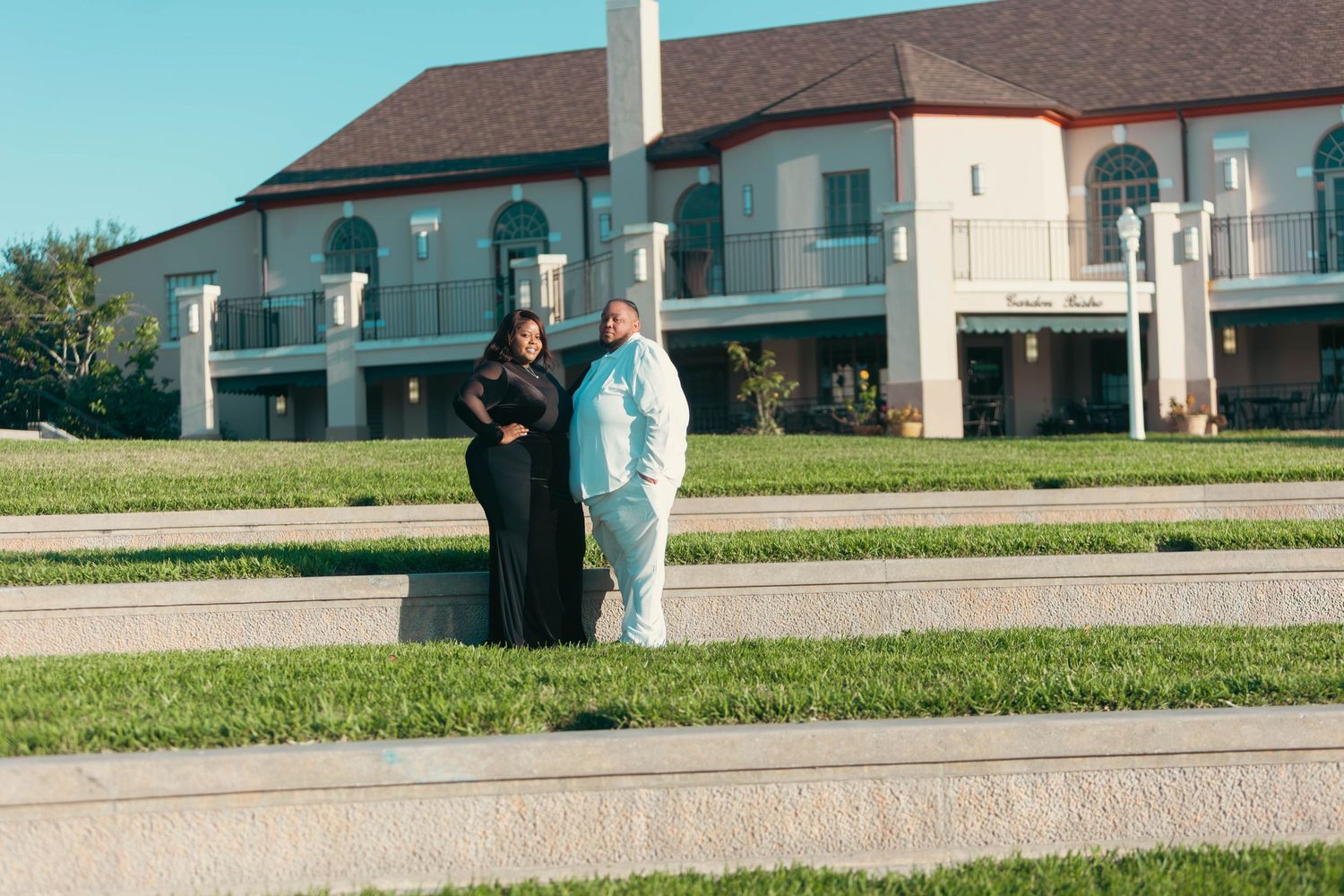 Couple standing side by side on a manicured lawn in front of a large white building outdoors.