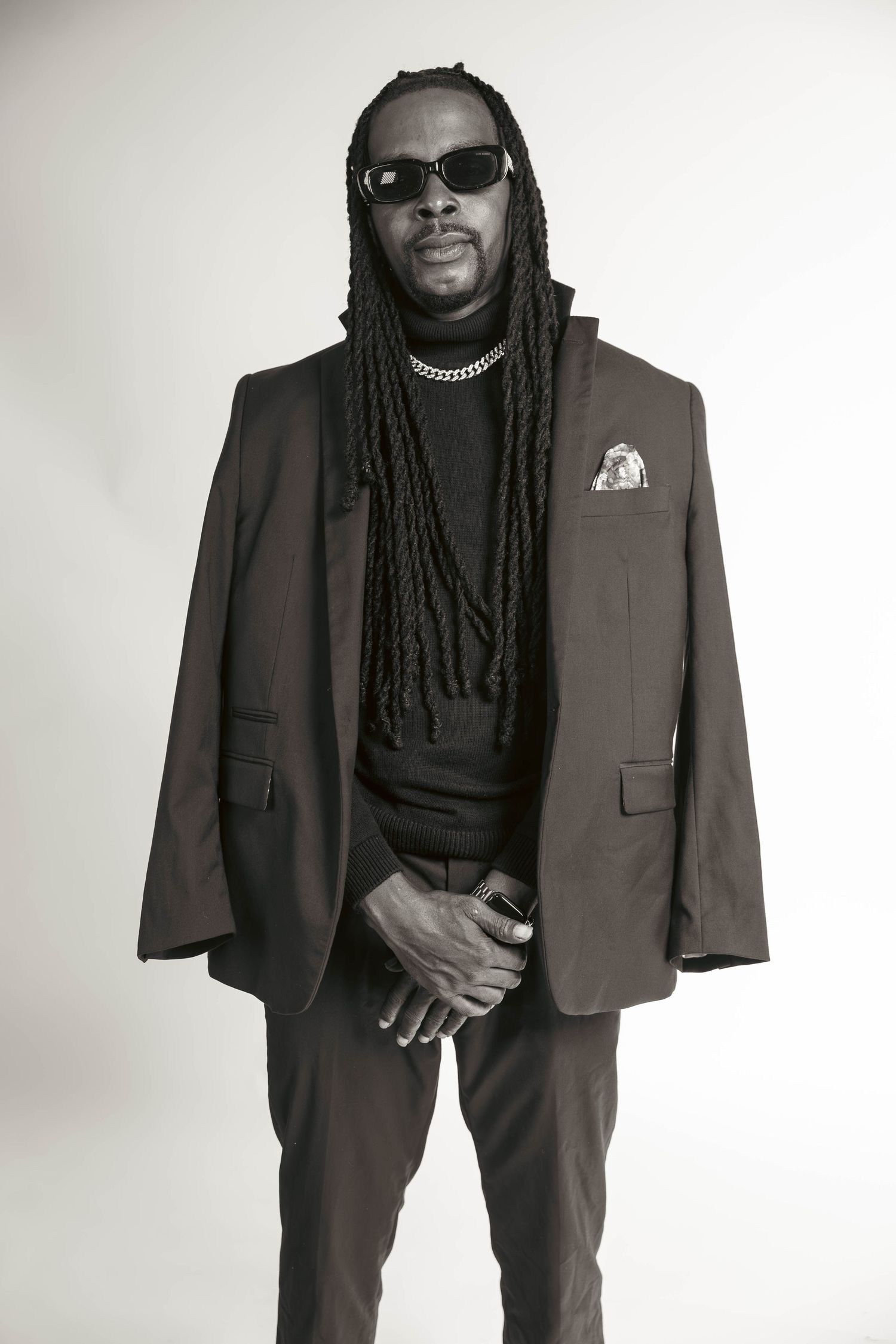 Black and white portrait of man in suit with long dreadlocks and sunglasses posing confidently.