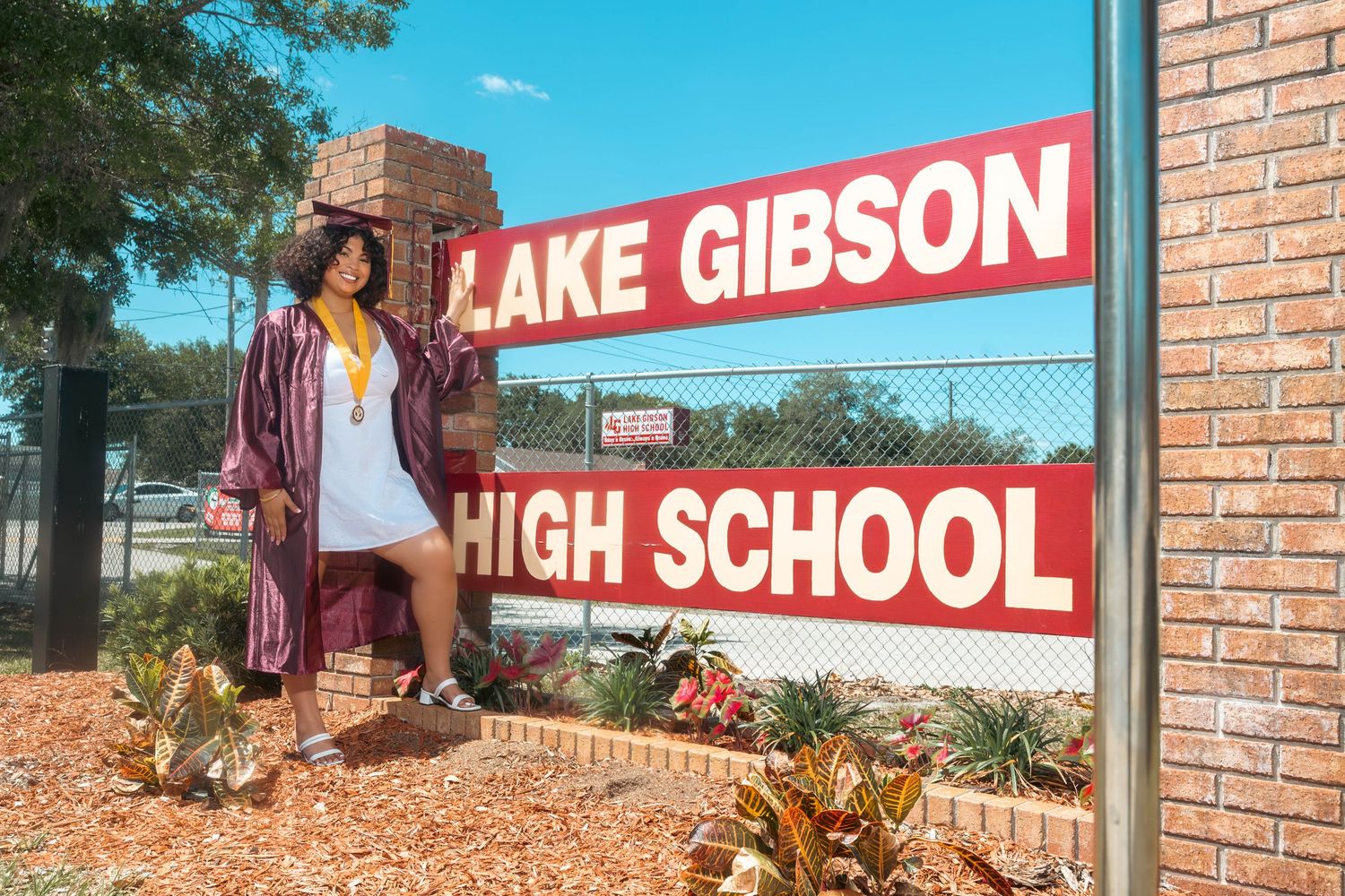 Graduate in maroon cap and gown poses proudly by Lake Gibson High School sign outdoors.