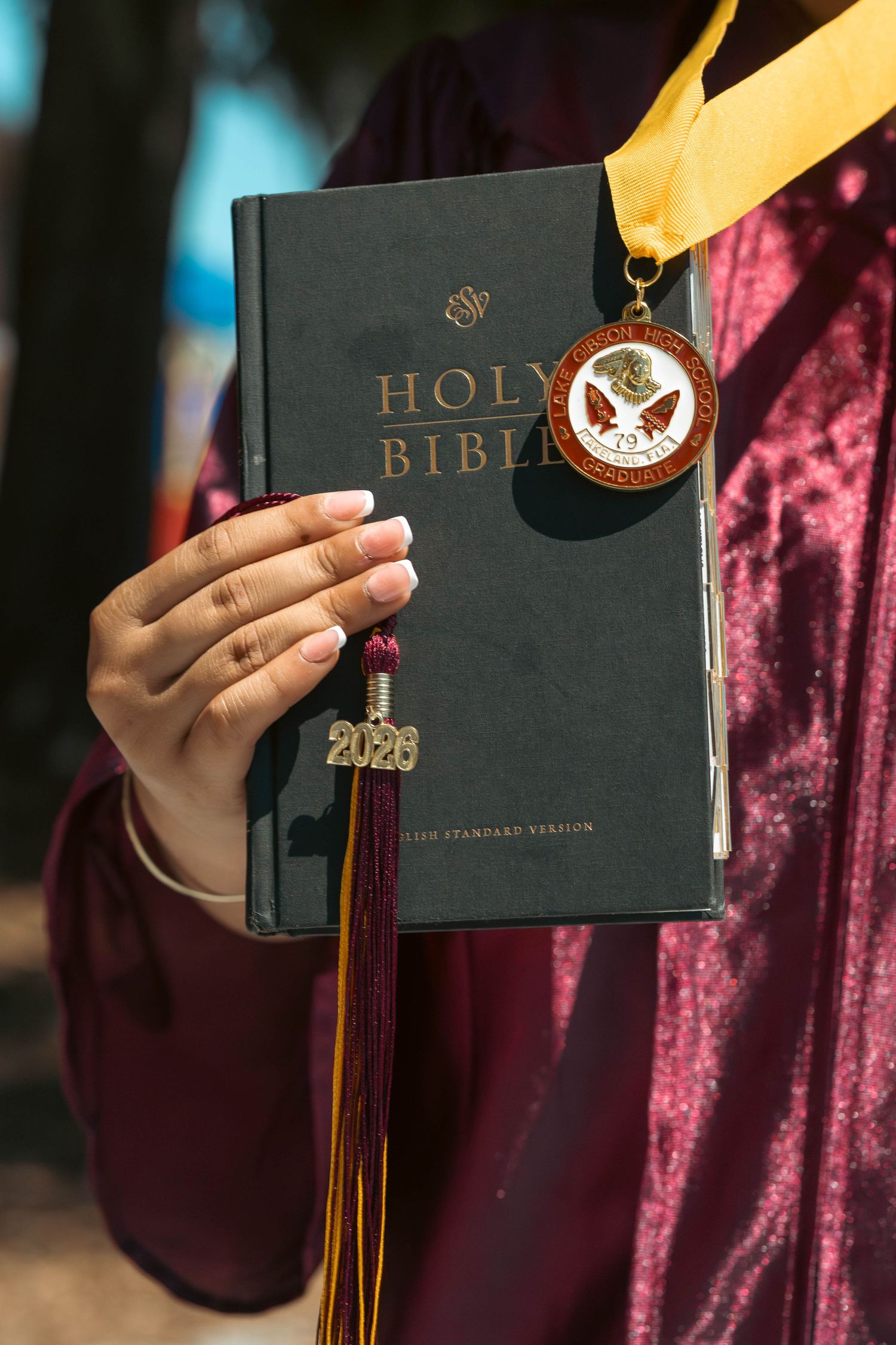 Close-up of graduate holding Holy Bible with maroon tassel charm and academic medal.