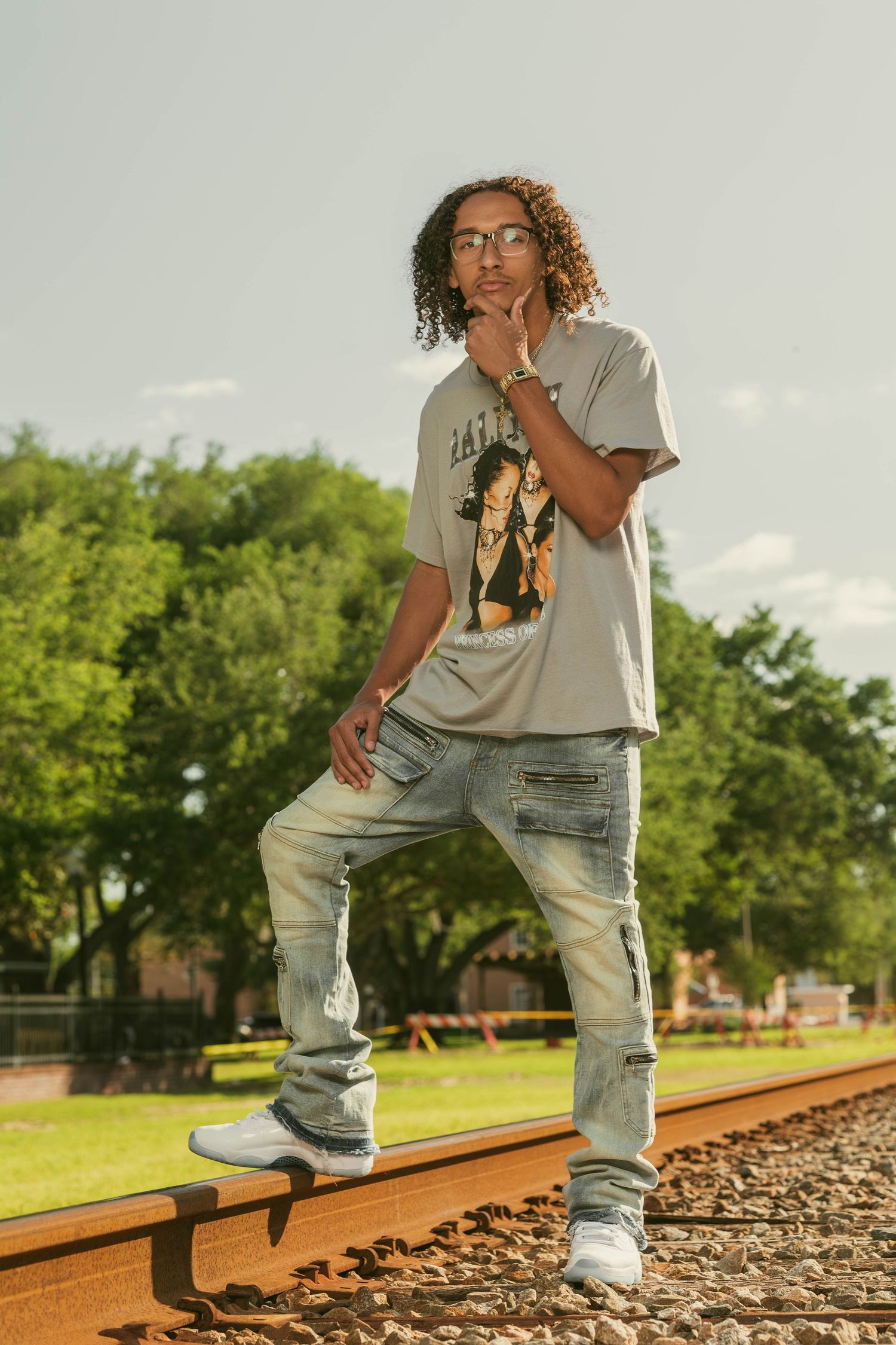 Young man with curly hair posing on railroad tracks in casual streetwear outfit.