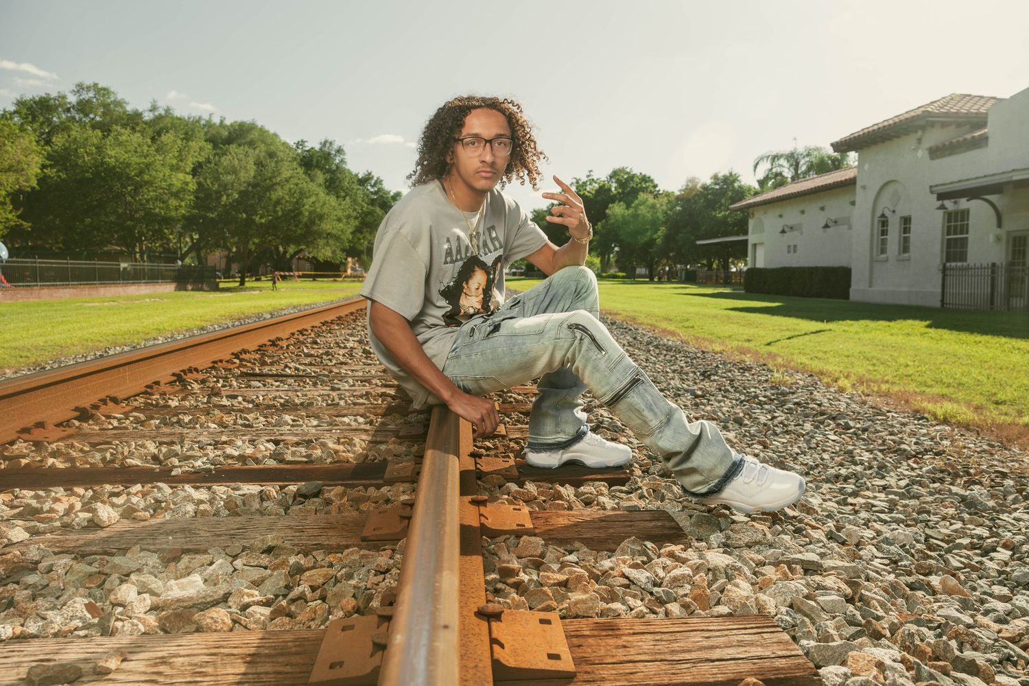 Young man with curly hair sitting on railroad tracks in casual streetwear outdoors.