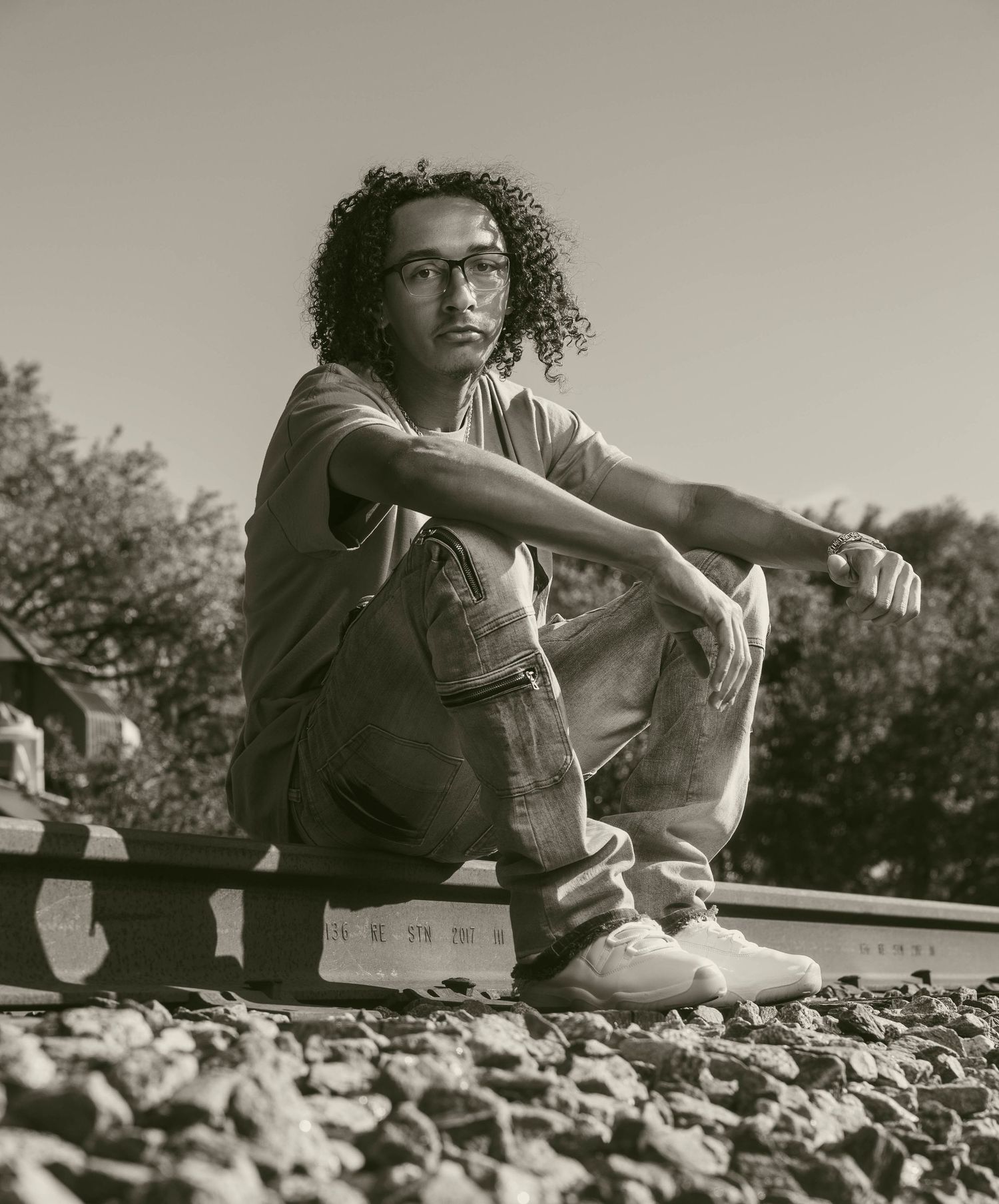 Young man with curly hair and glasses sits on railroad tracks in a moody black and white portrait photo.