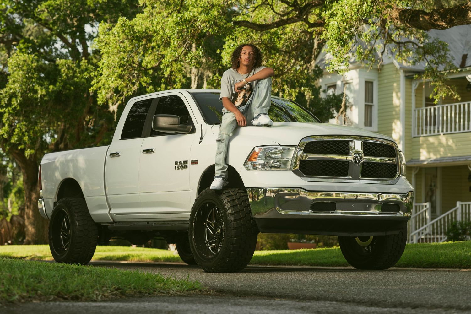 Person sitting on hood of lifted white Ram 1500 truck parked on residential street surrounded by trees.