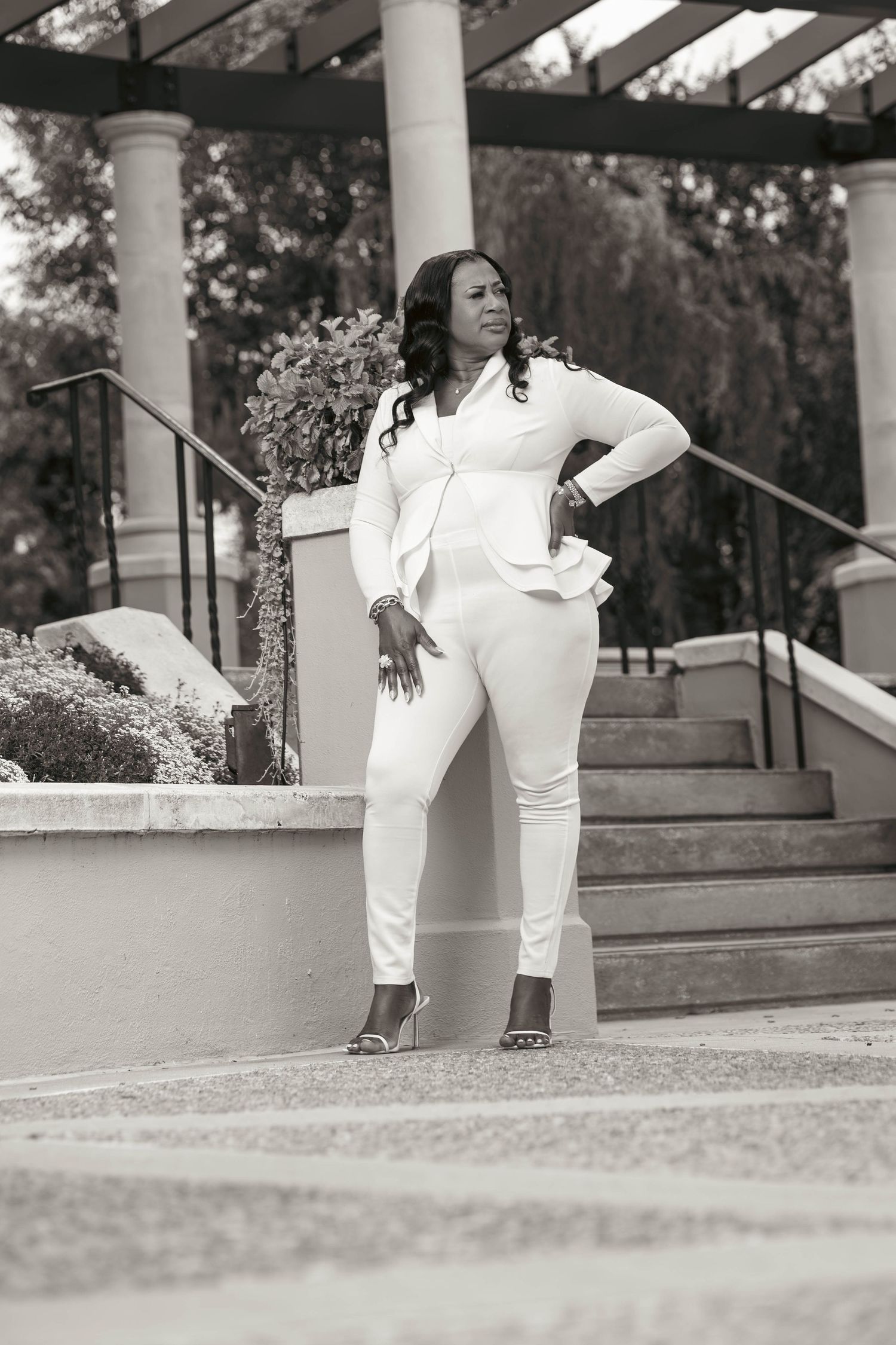 Confident Black woman in white pantsuit strikes a bold pose on outdoor staircase in black and white photo.
