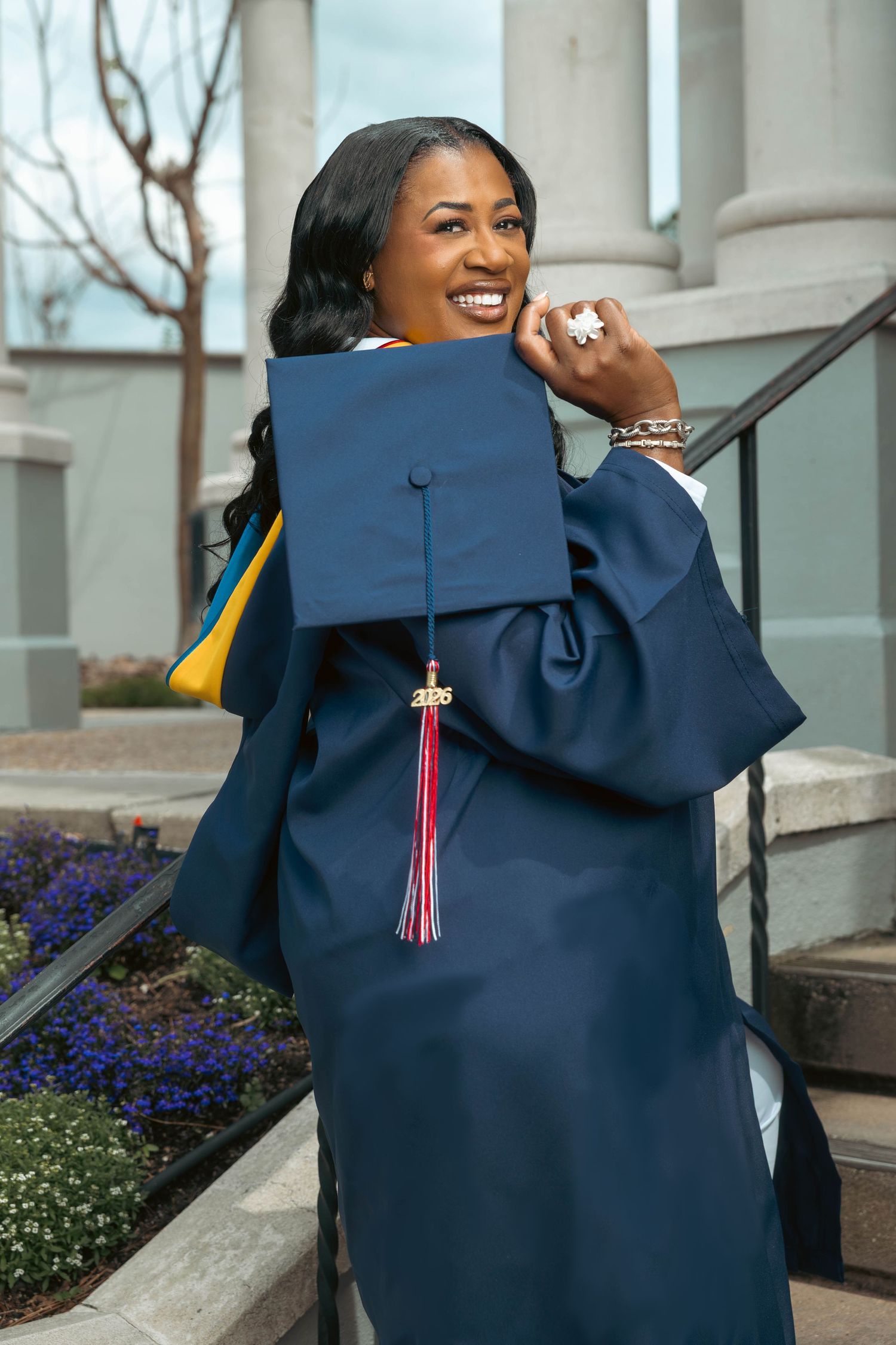 Smiling Black woman in navy graduation cap and gown holds mortarboard on outdoor staircase steps.