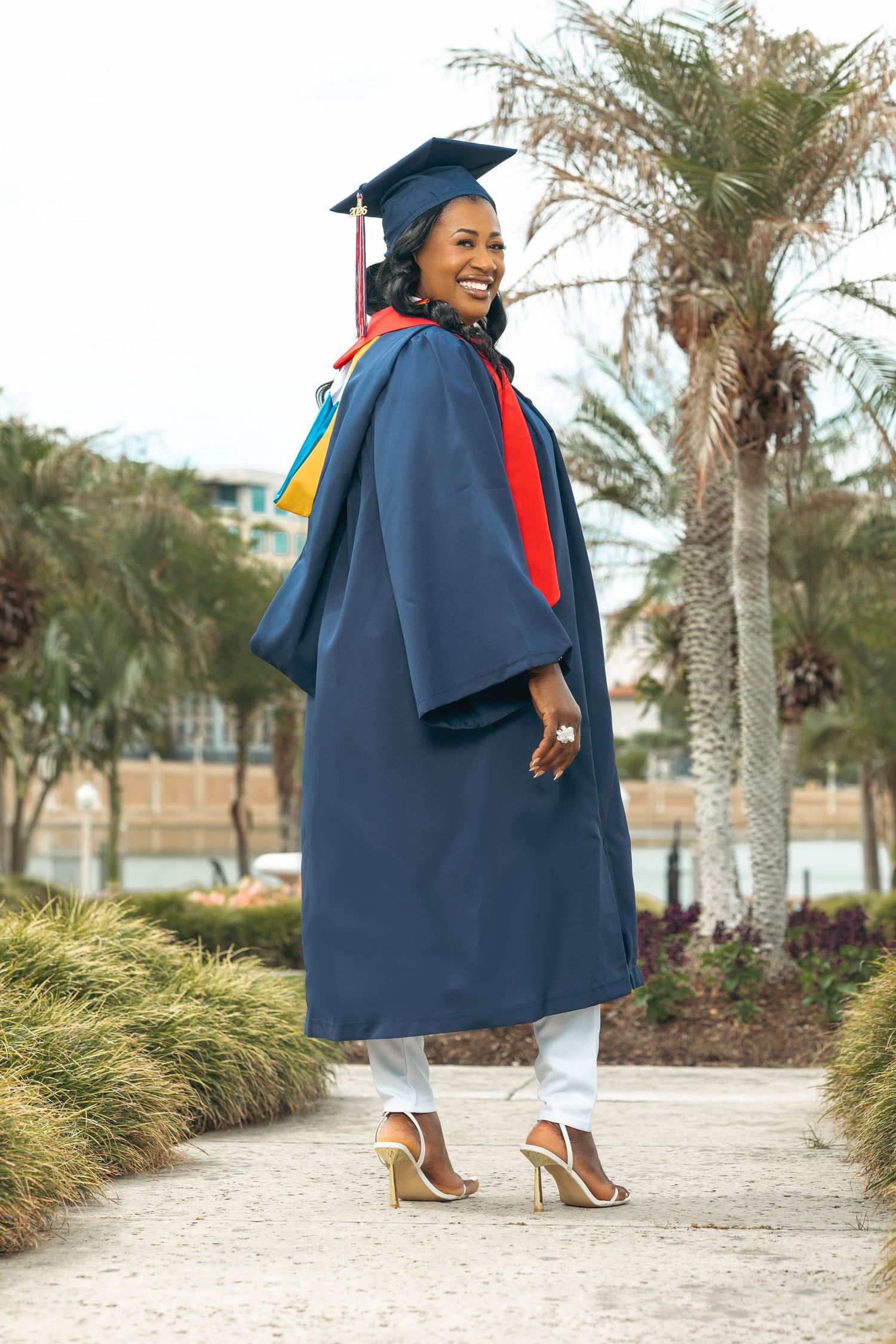 Smiling graduate in navy cap and gown with colorful stoles poses outdoors near palm trees.