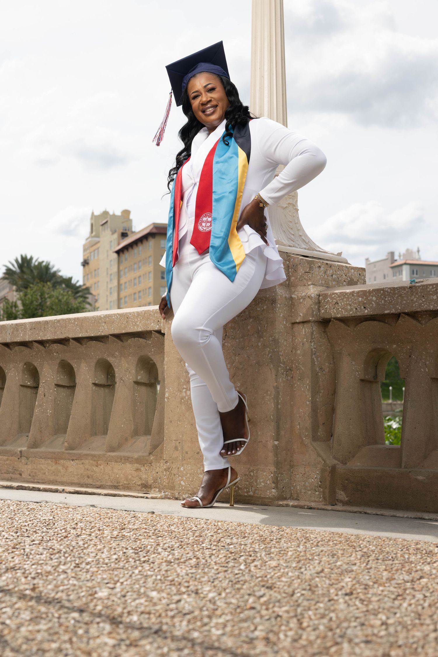 Graduate in white outfit with colorful stoles and cap poses confidently by stone balustrade.