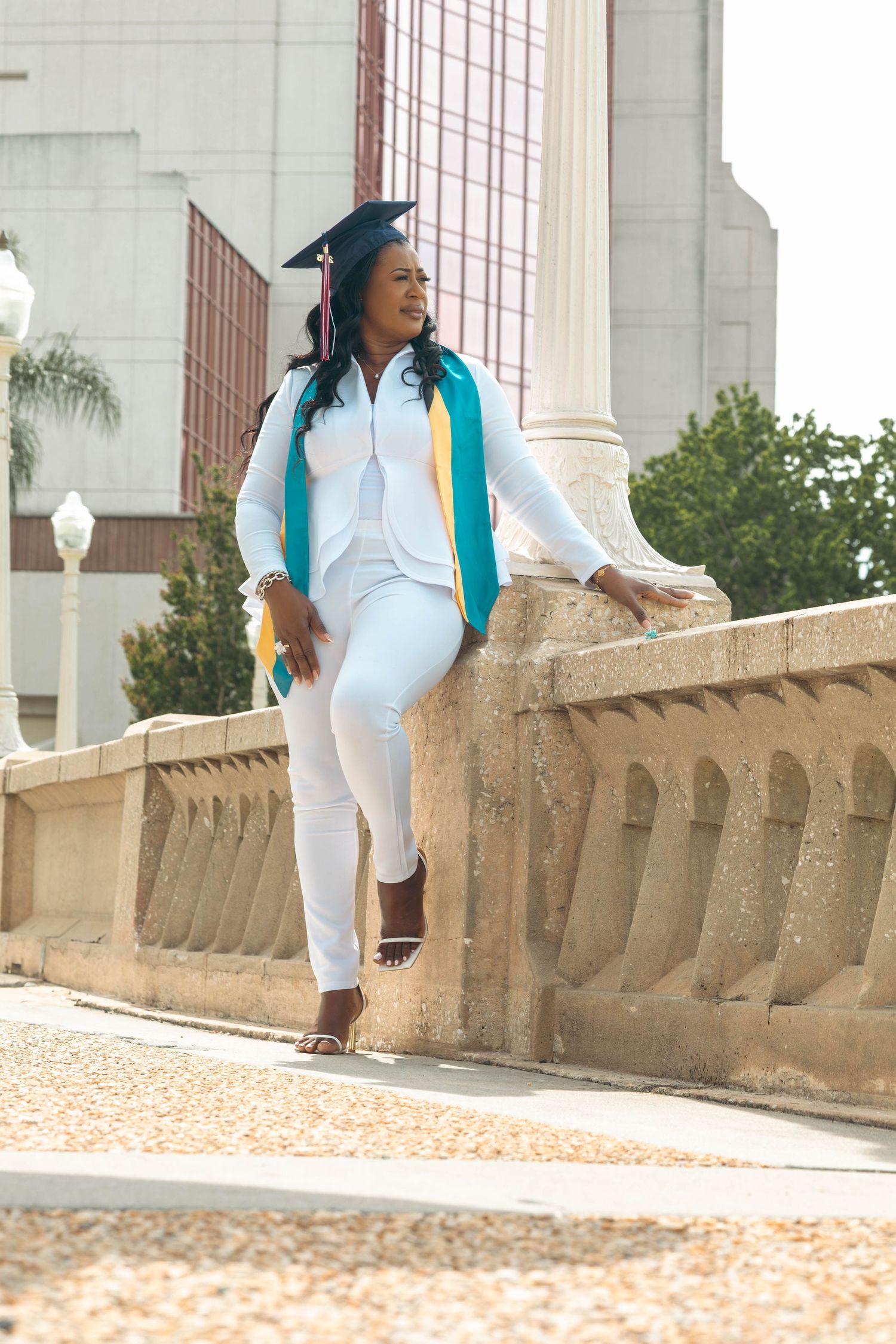Graduate in white outfit with teal stole leans against stone railing near a column outdoors.
