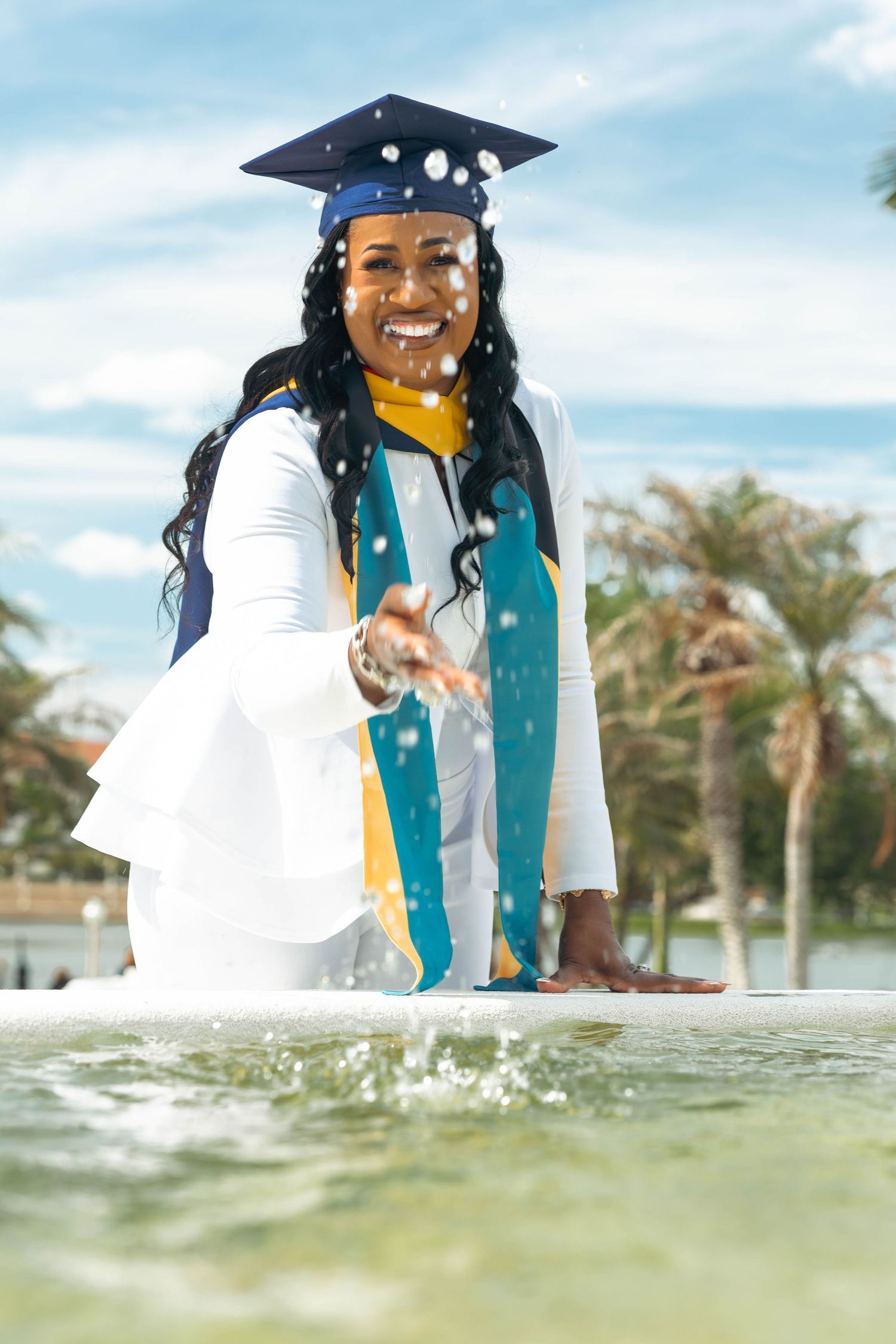 Joyful graduate in white outfit and cap splashes water in a fountain surrounded by palm trees.