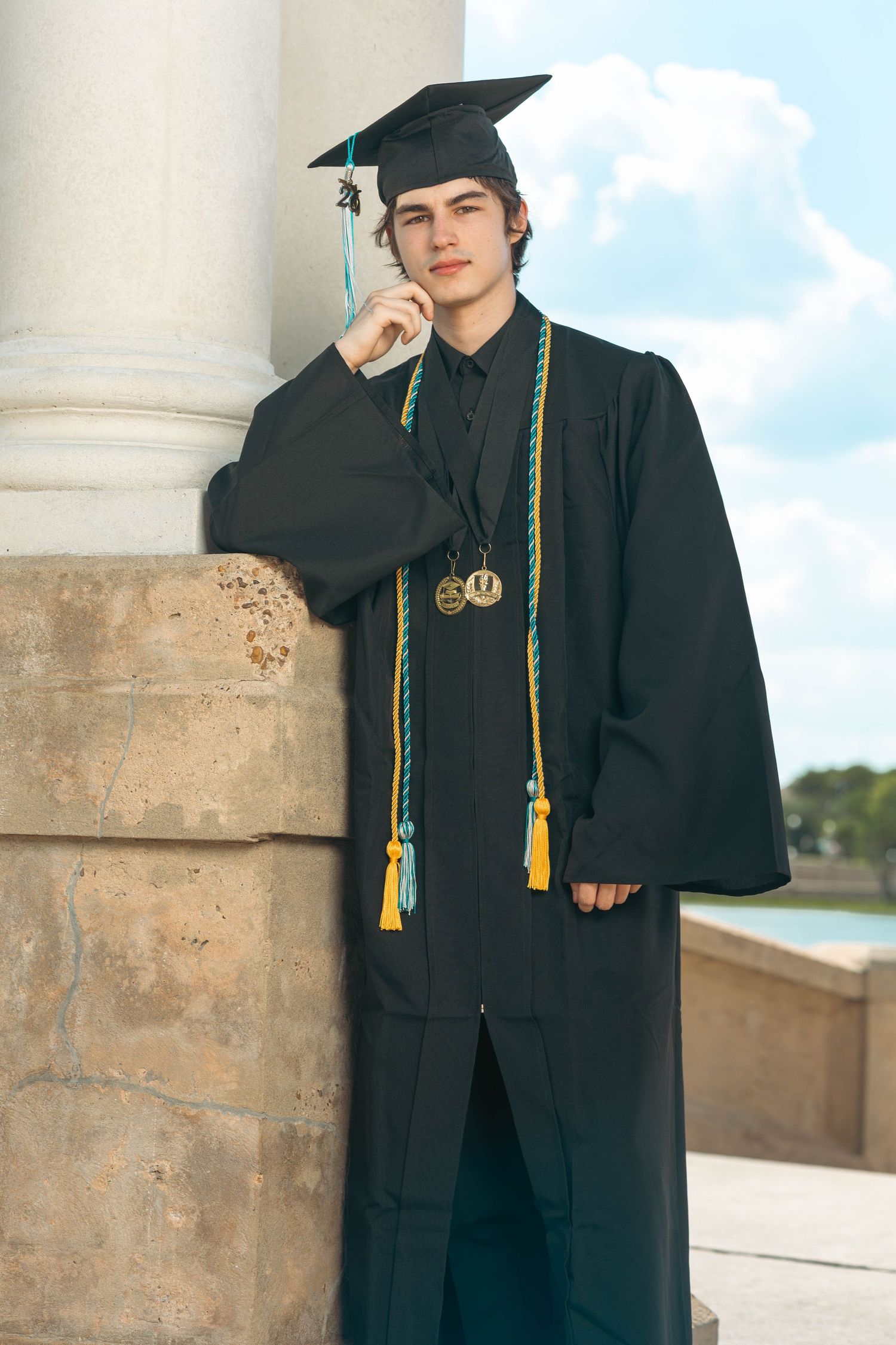 Young male graduate in cap and gown leaning against stone column outdoors.