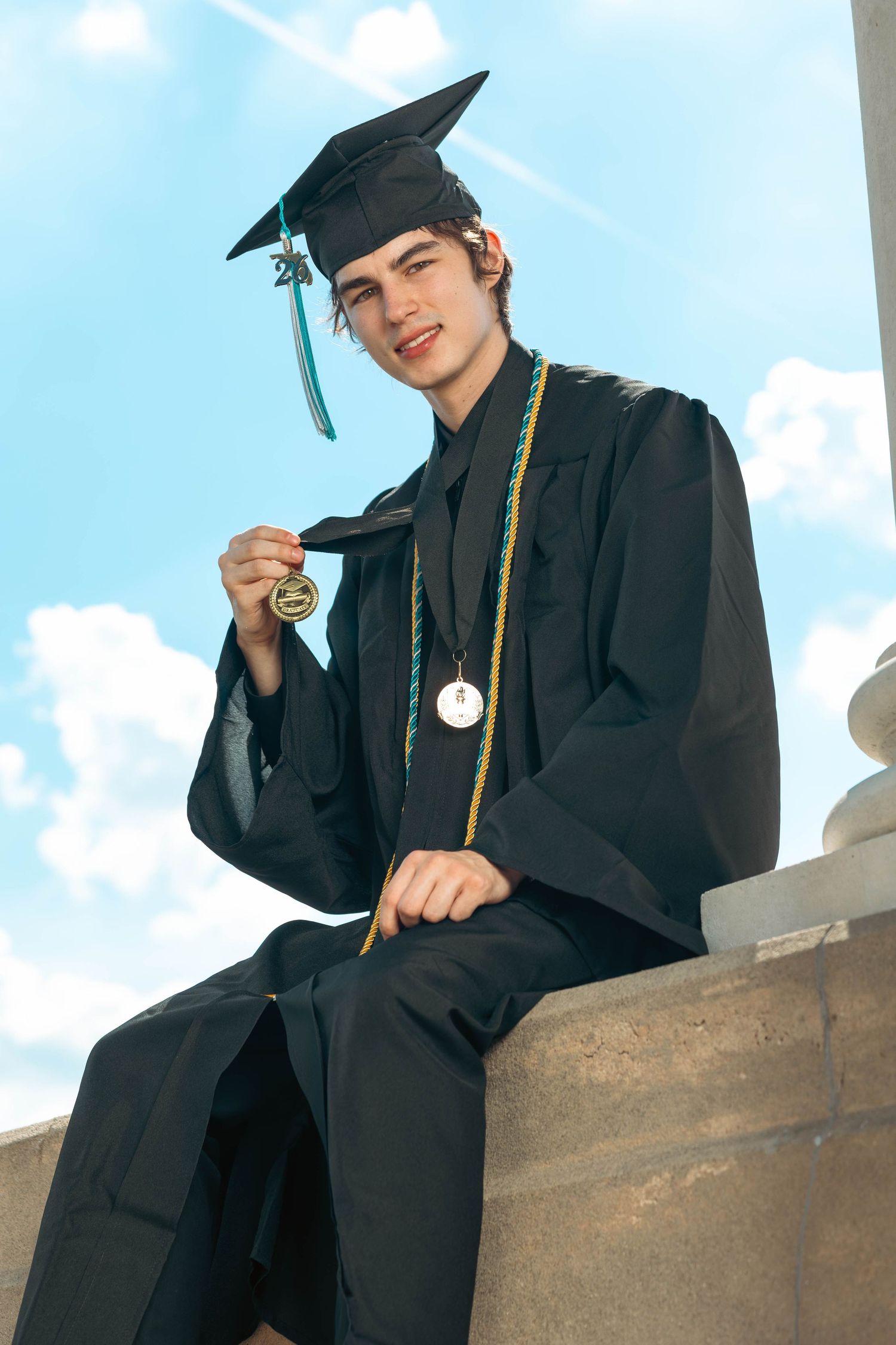 Smiling male graduate in cap and gown holding medal while seated on stone ledge.