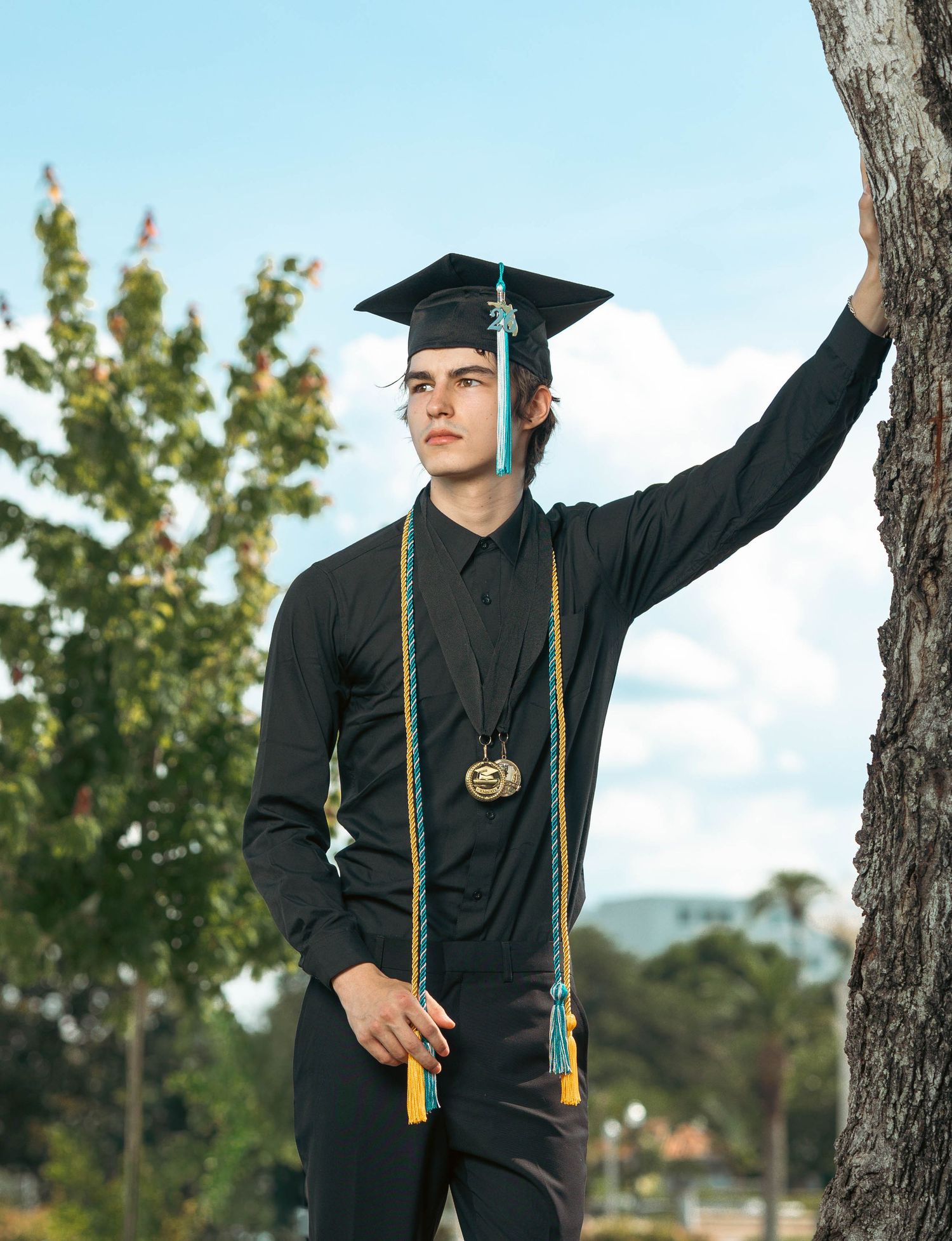 Male graduate in cap and gown leaning against tree in outdoor setting.