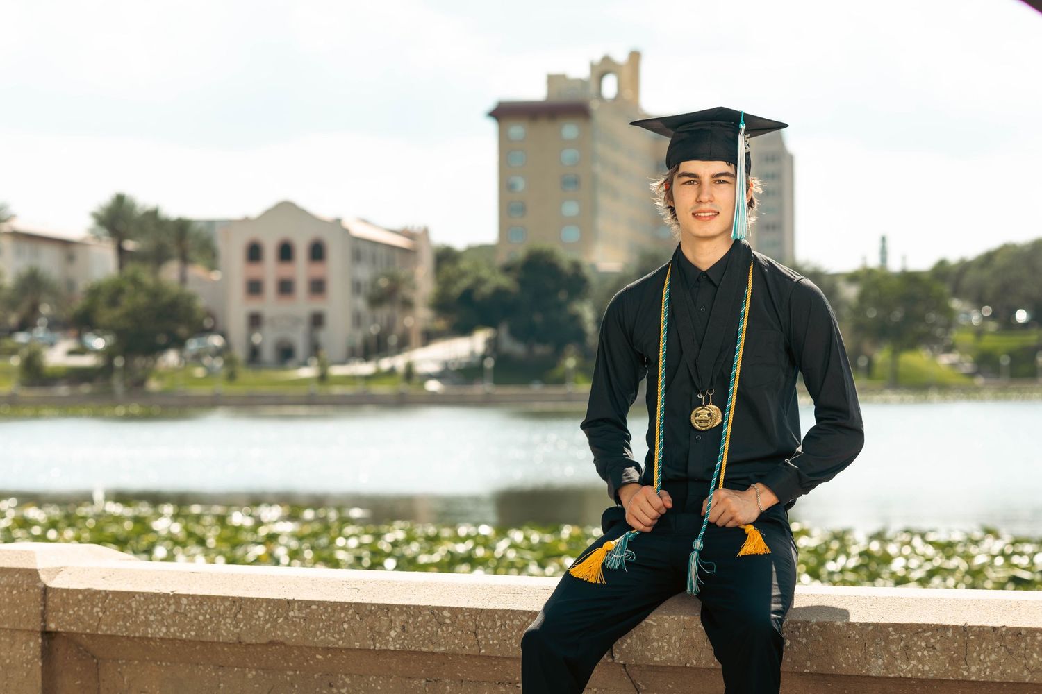 Male graduate in cap and gown sitting on ledge with scenic waterfront background.