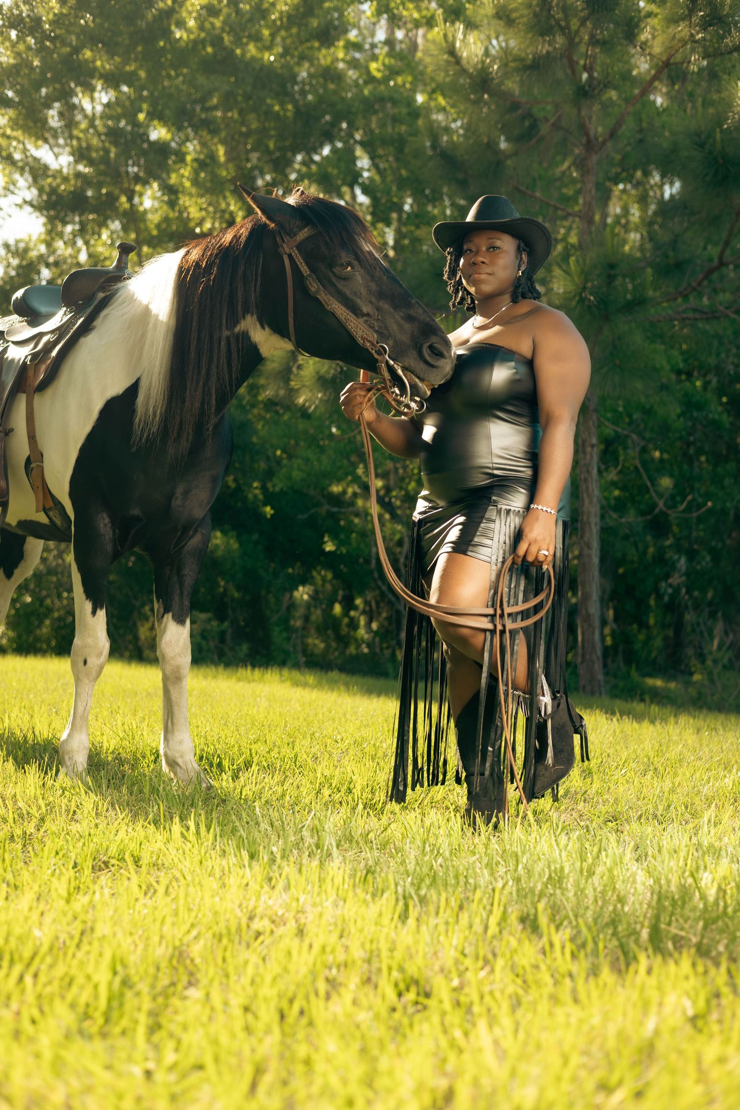Black woman in leather outfit and cowboy hat stands beside paint horse in sunny green field.